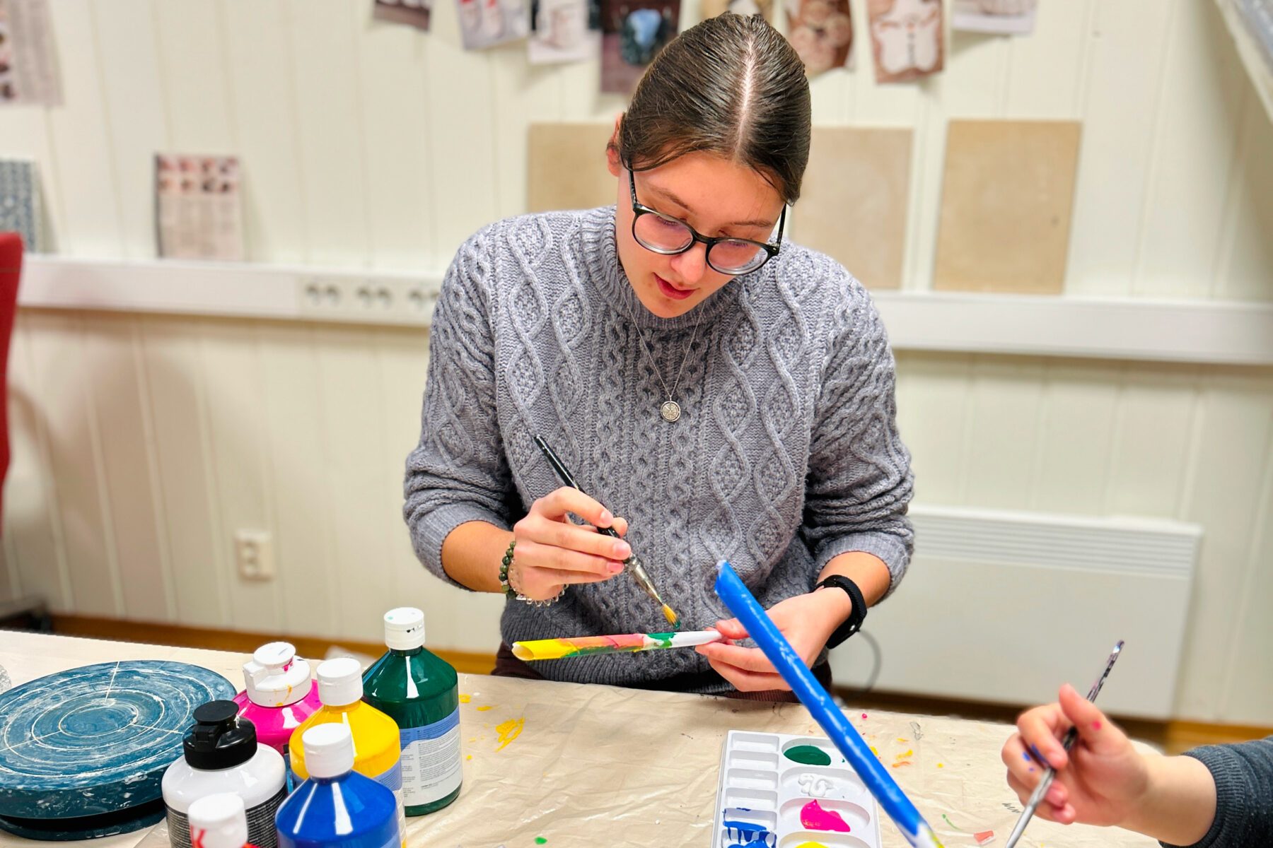ASF-Freiwillige in grauem Zopfmusterpullover mit Brille bemalt ein buntes Holzstück; Farben, Pinsel und Palette auf Tisch.