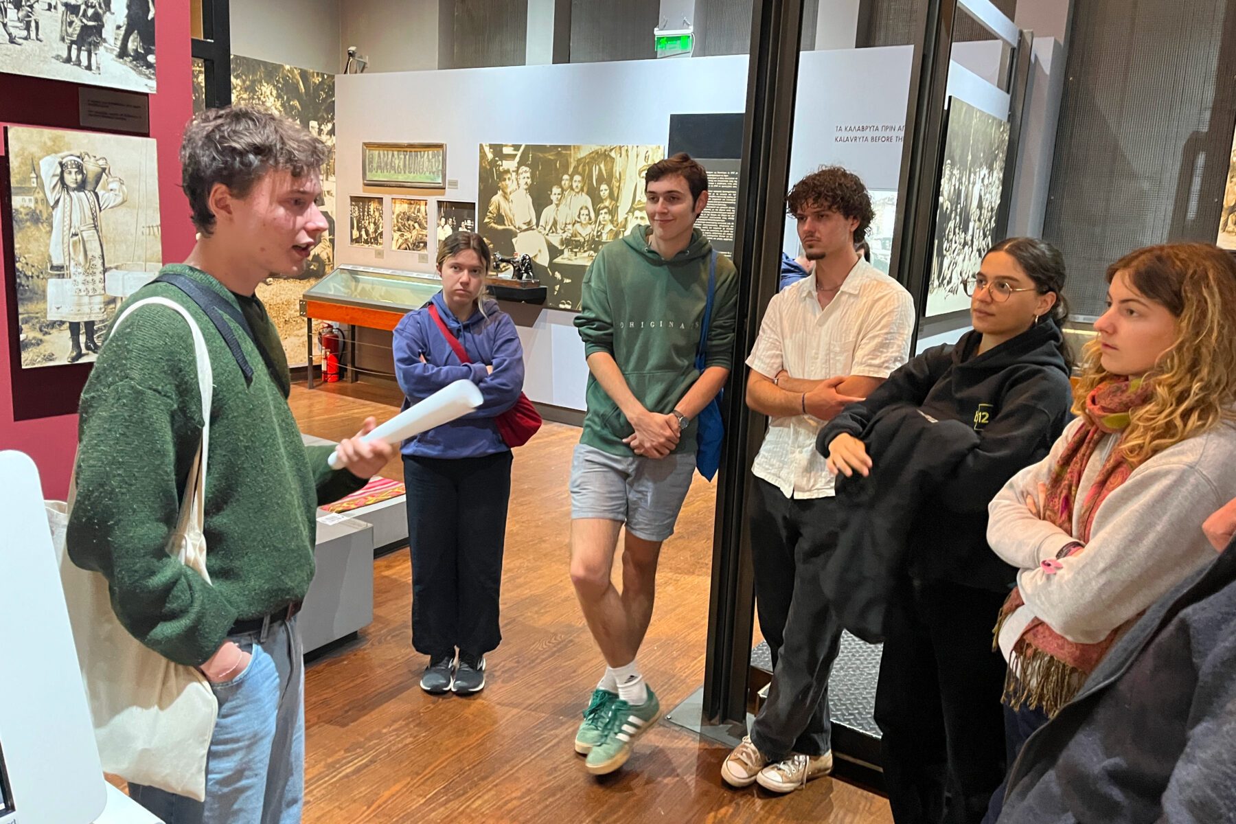 ASF‑Freiwilliger spricht mit Jugendlichen im Holocaust‑Museum Kalavryta, Gruppe vor Ausstellung, er hält ein gerolltes Papier