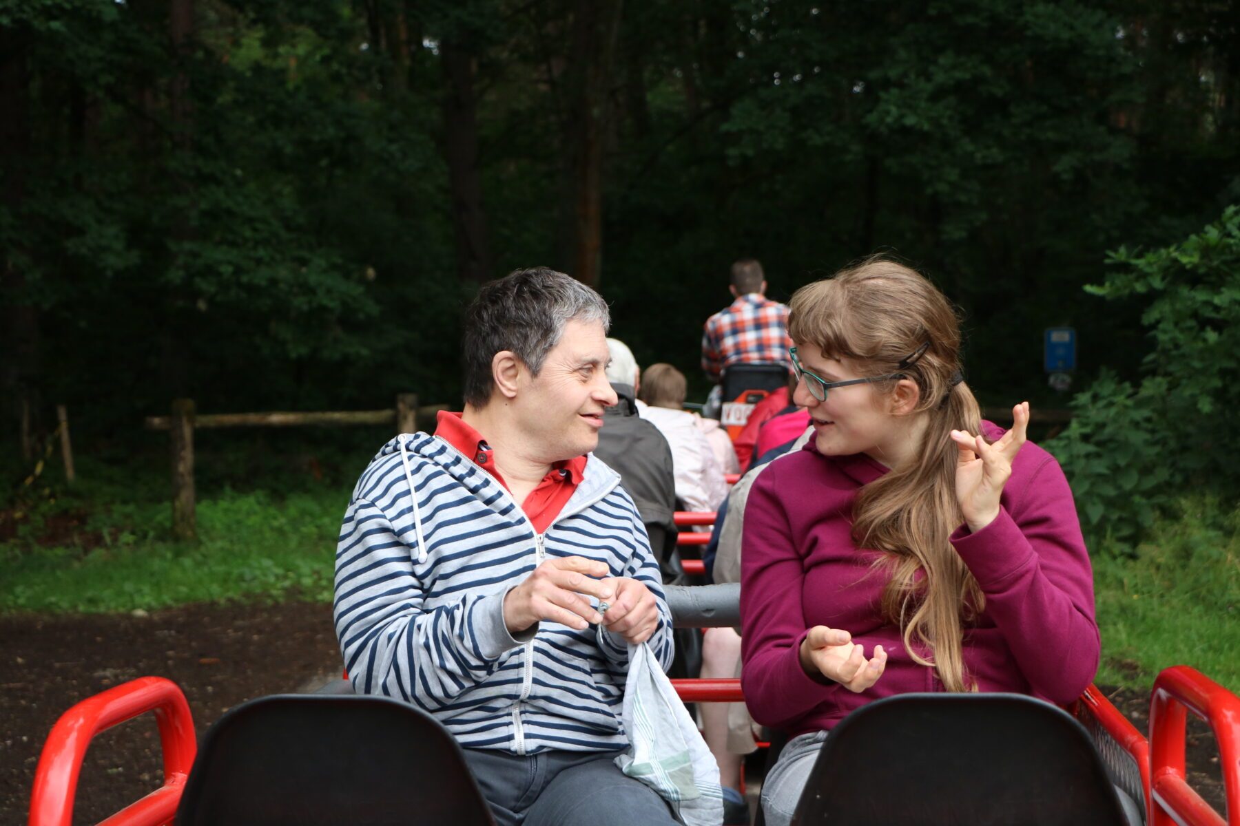 Zwei Frauen sitzen in roten Sitzen eines Ausflugszugs im Wald; eine gestreift, die andere lila, sie gestikulieren.