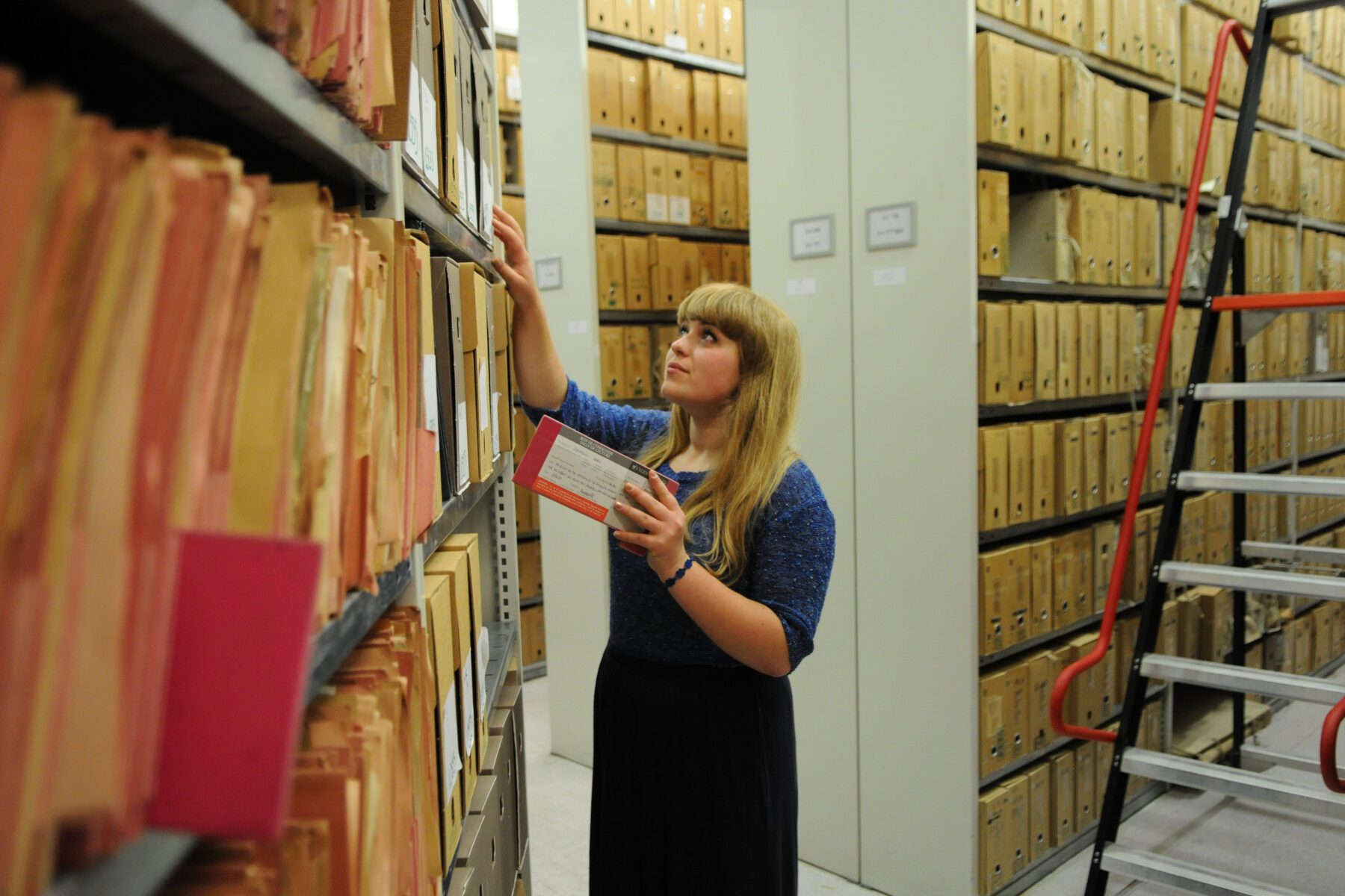 Freiwillige mit verpixeltem Gesicht greift in Archivregal nach Box, Reihen voller Kartons, rote Leiter rechts.