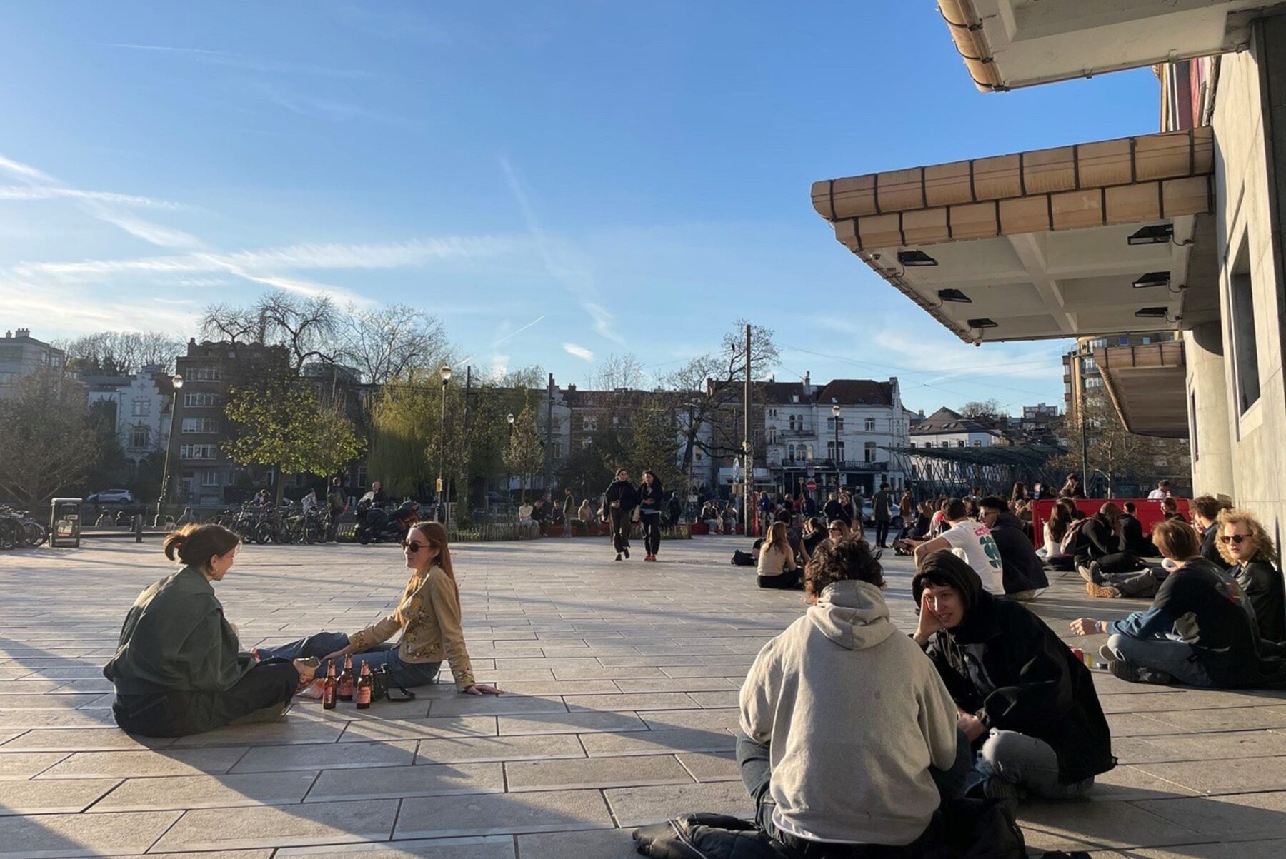 ASF-Freiwillige sitzen in Gruppen auf sonnigem Platz in Brüssel, plaudernd; Flaschen am Boden, Gebäudevordach rechts.