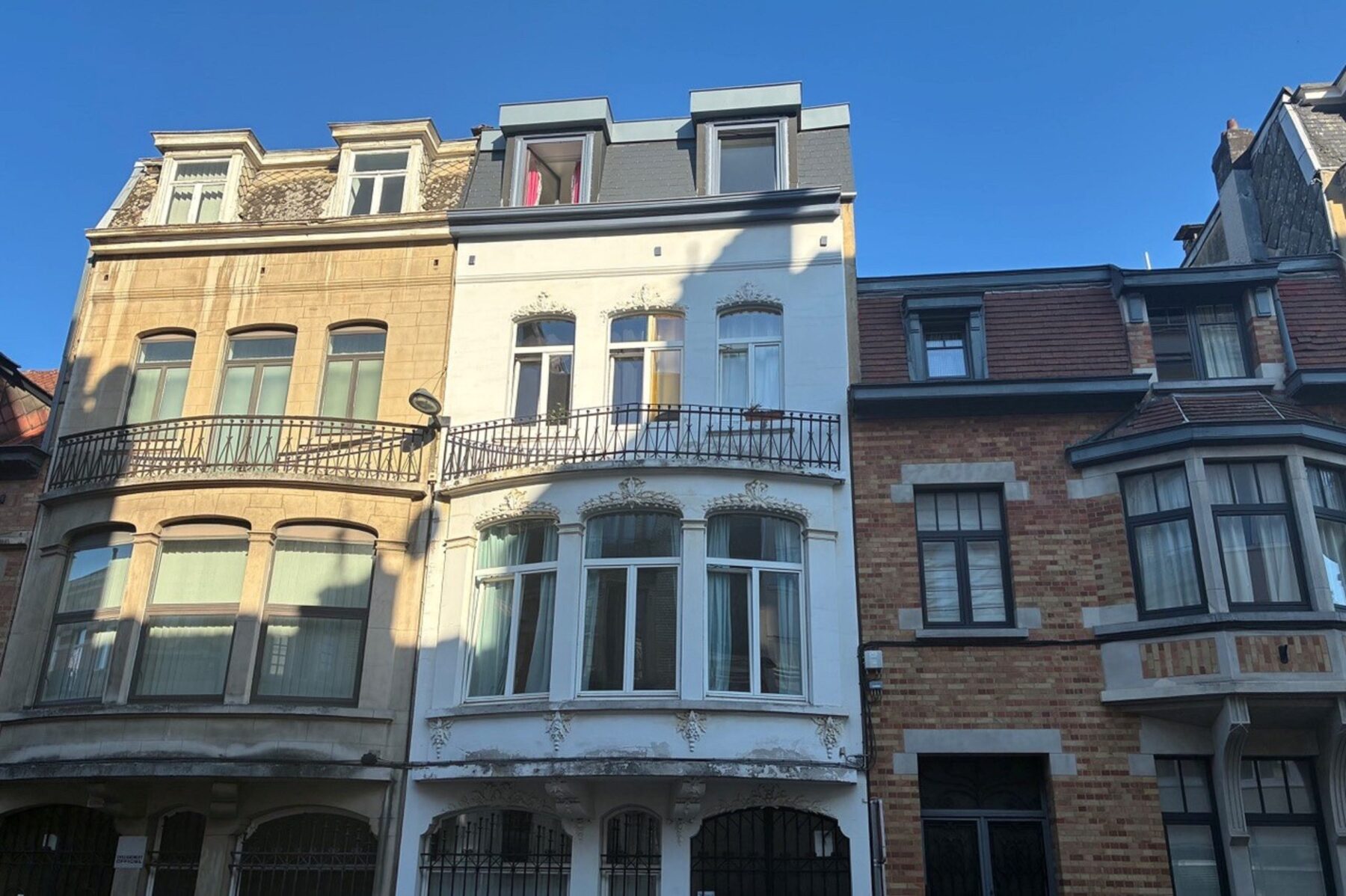 Drei Reihenhäuser in Brüssel: mittiges weißes Haus mit Erker und Balkon, links beige, rechts Ziegelbau, blauer Himmel.