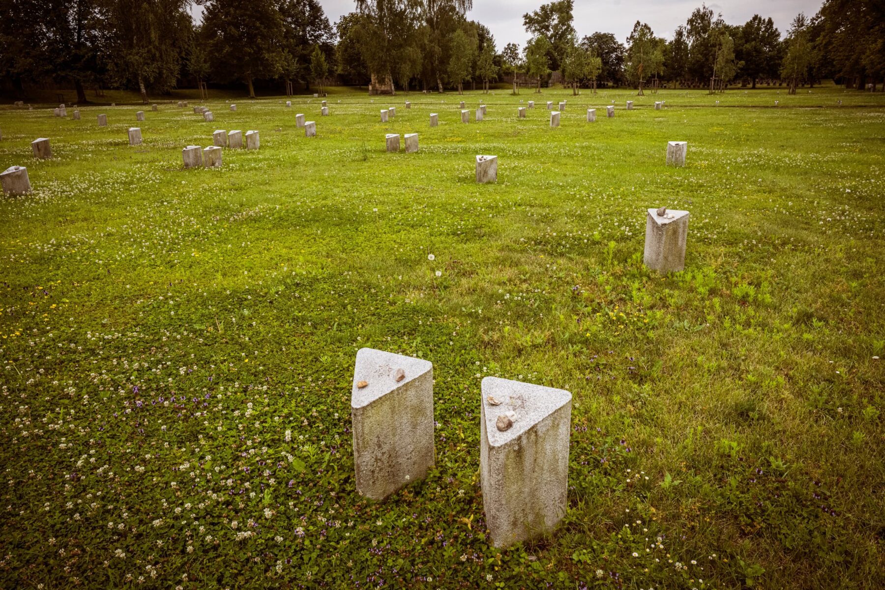 Gedenkstätte Terezín: Wiese mit Reihen dreieckiger Steinsäulen, zwei Stelen mit kleinen Gedenksteinen im Vordergrund