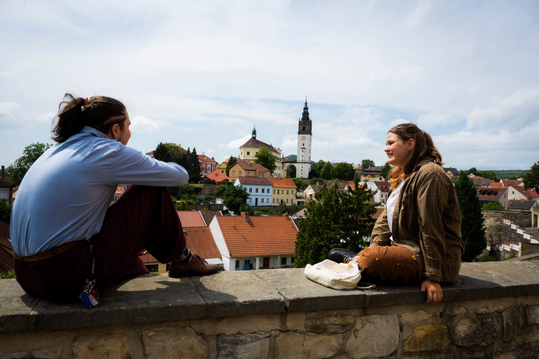 Zwei Freiwillige sitzen auf einer Steinmauer und blicken auf Terezín mit Kirchturm und roten Dächern.