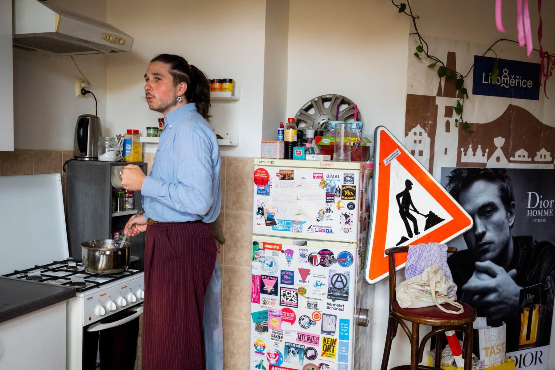 Junger Freiwilliger in hellblauem Hemd rührt Topf am Herd, aufgeklebter Sticker-Kühlschrank, Baustellenschild und Dior-Poster.