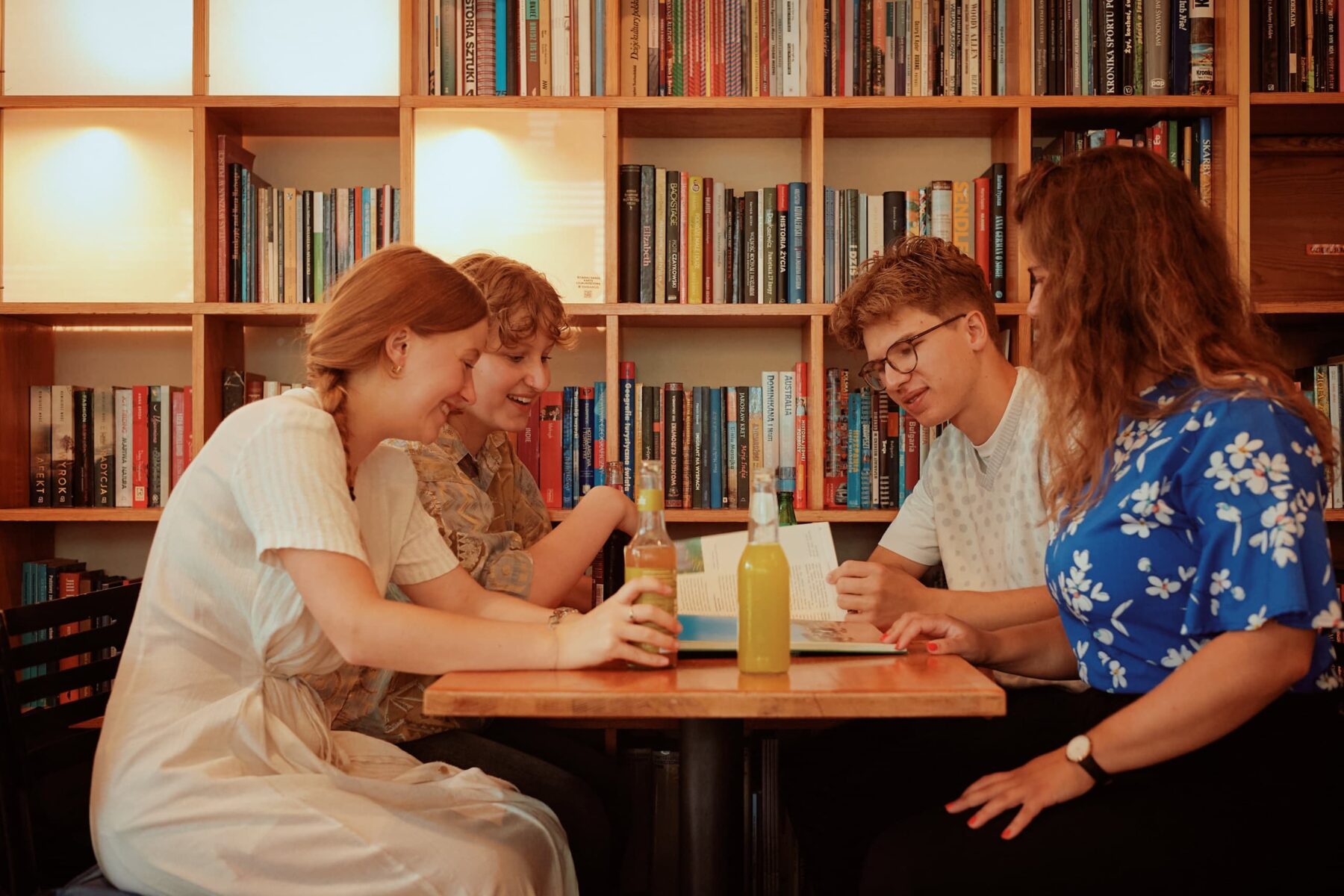 Vier ASF-Freiwillige sitzen an einem Tisch vor Bücherregal, lesen ein Buch, trinken gelbe Getränke und lachen.