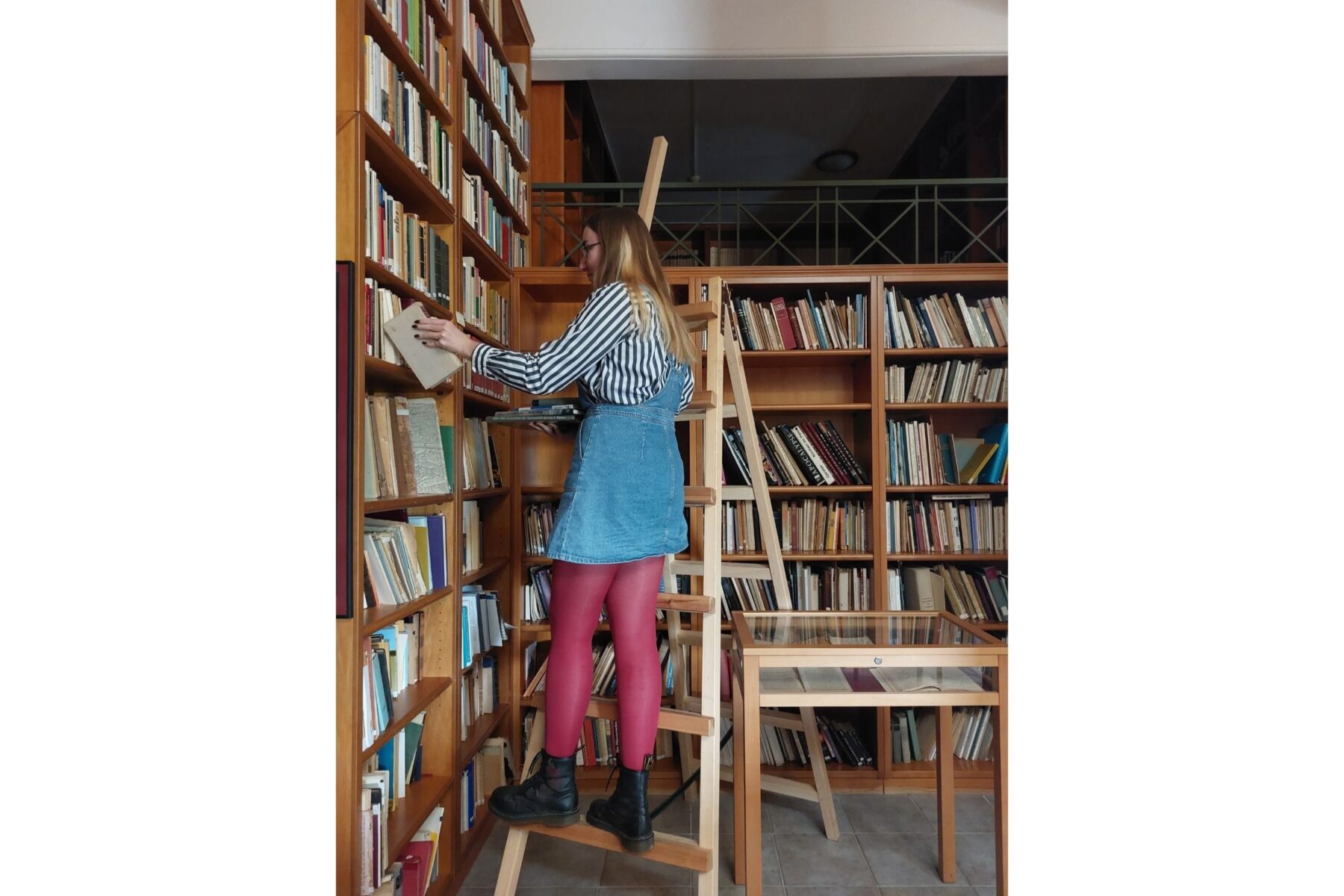 Freiwillige auf Leiter in Bibliothek der Orthodoxen Akademie Kreta, zieht Buch aus Regal, Jeanskleid und pinke Strumpfhose.