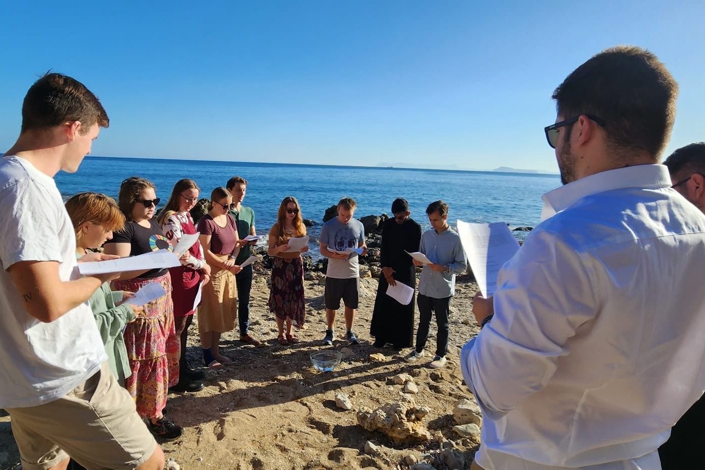 Freiwillige der orthodoxen Akademie auf Kreta stehen im Kreis am Meer, halten Texte während einer Zeremonie.