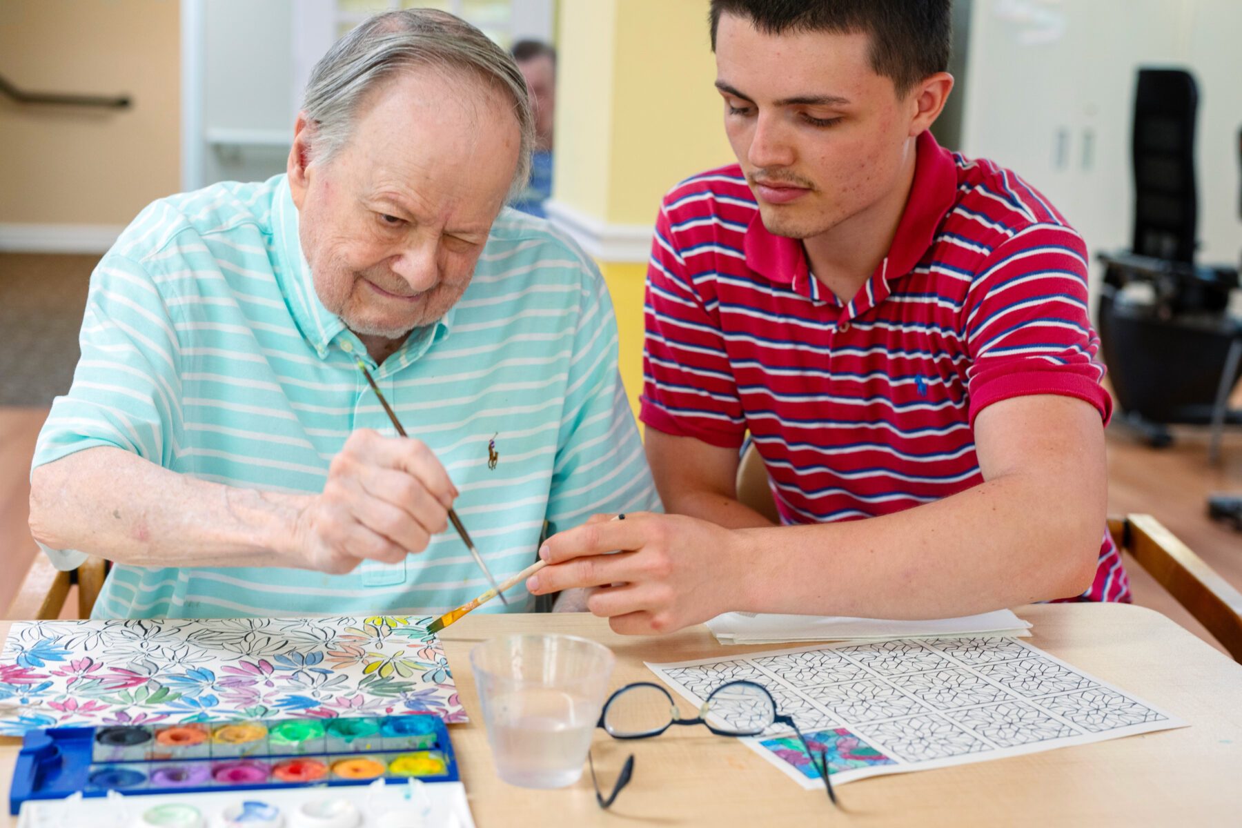 Freiwilliger und Senior malen zusammen, Pinsel, Aquarellkasten, Wasserglas und Ausmalvorlagen auf dem Tisch.