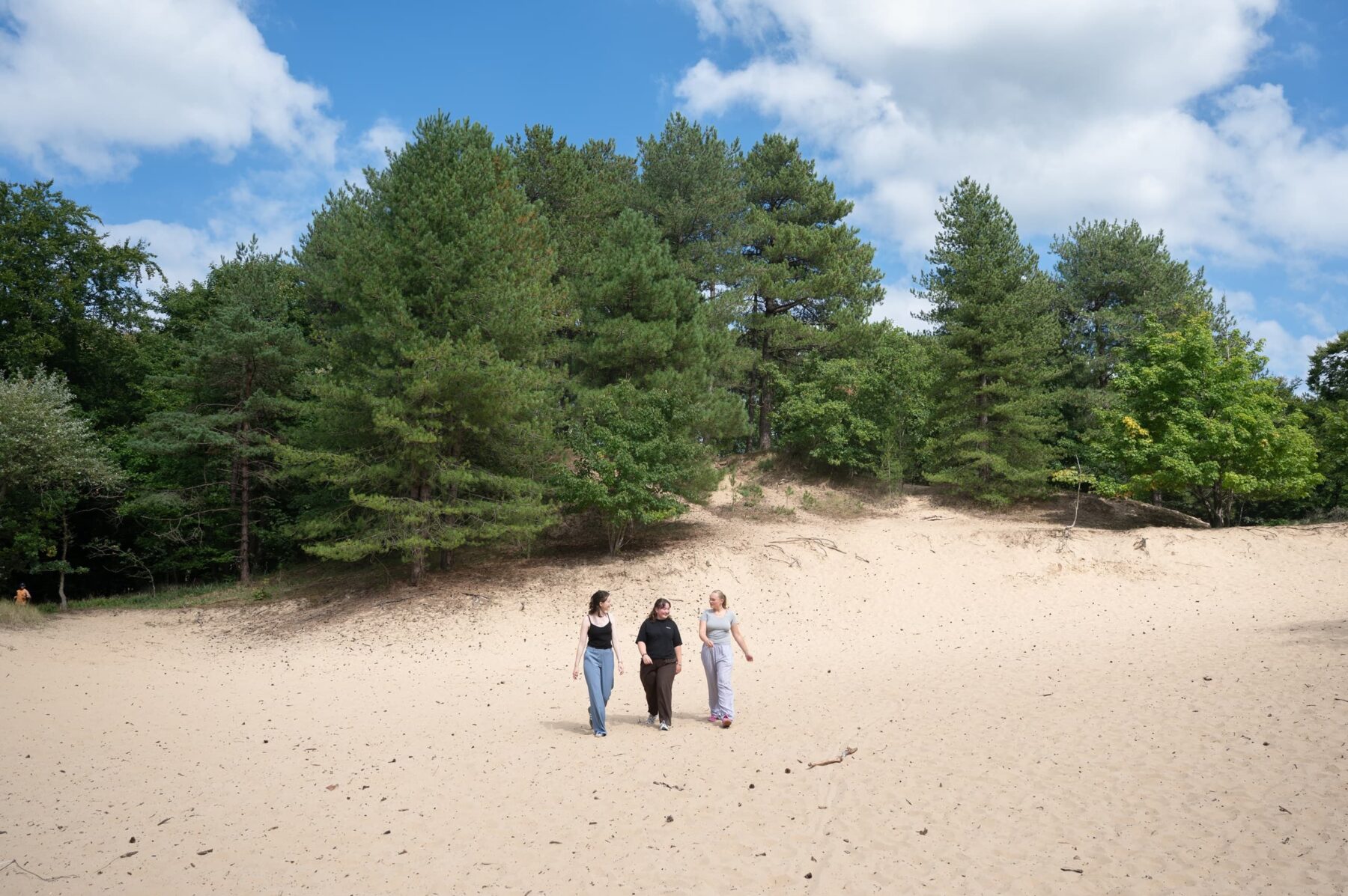 Drei Freiwillige laufen über helle Dünen, hinter ihnen Kiefernwald und blauer Himmel am Strand von Bloemendaal.