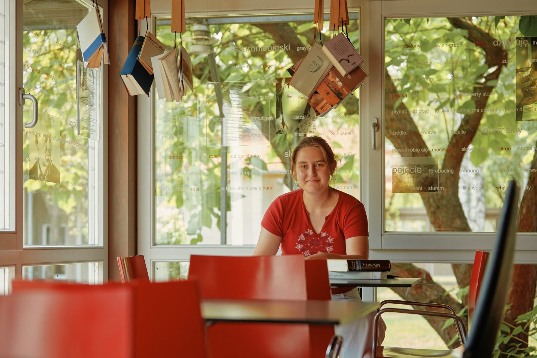 Frau in rotem Shirt sitzt an Tisch mit Büchern im IJBS Oświęcim, Polen 2024. Grünen Garten im Hintergrund.