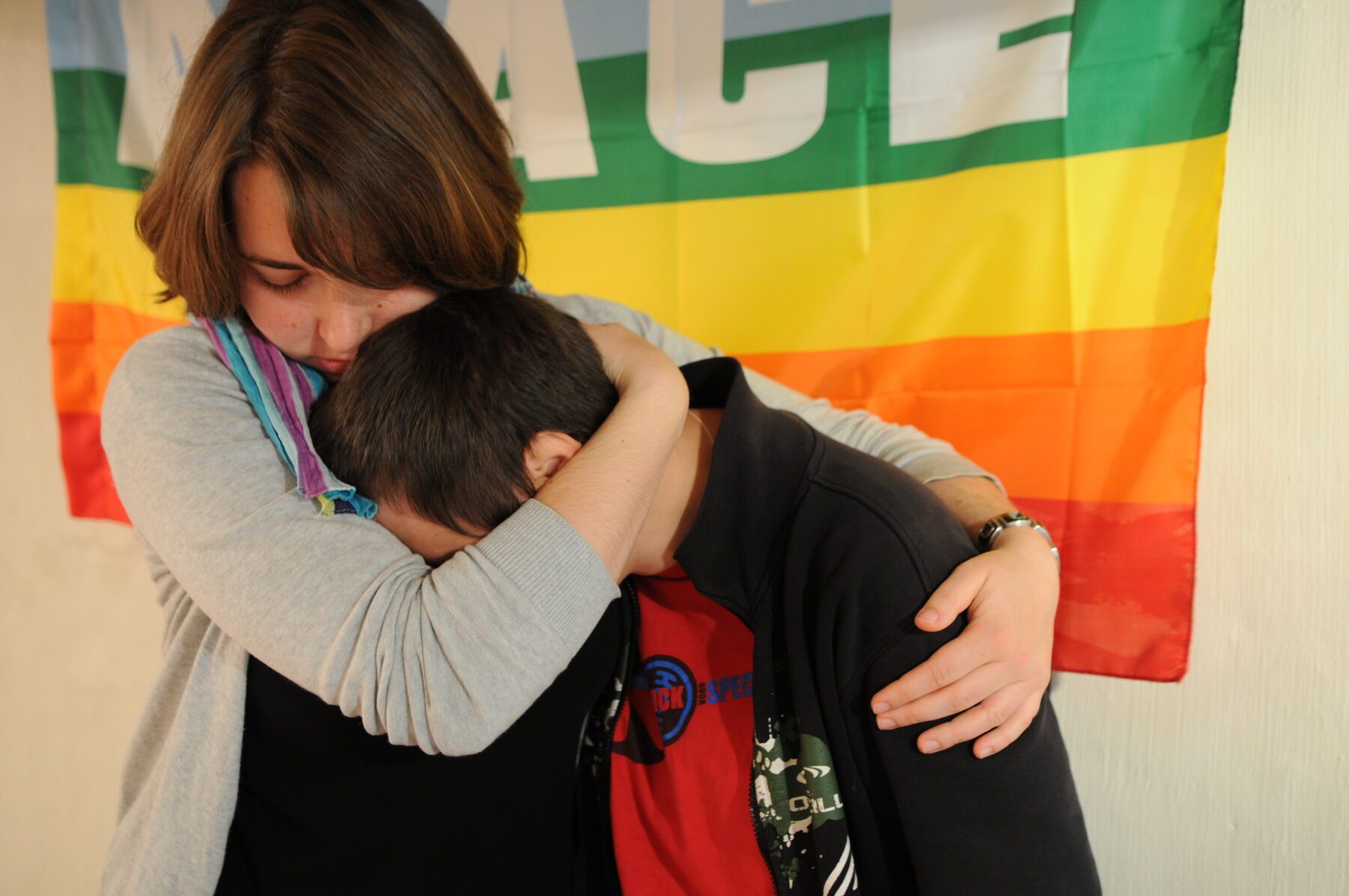 Freiwillige umarmt einen Jungen eng vor einer bunten Regenbogenflagge