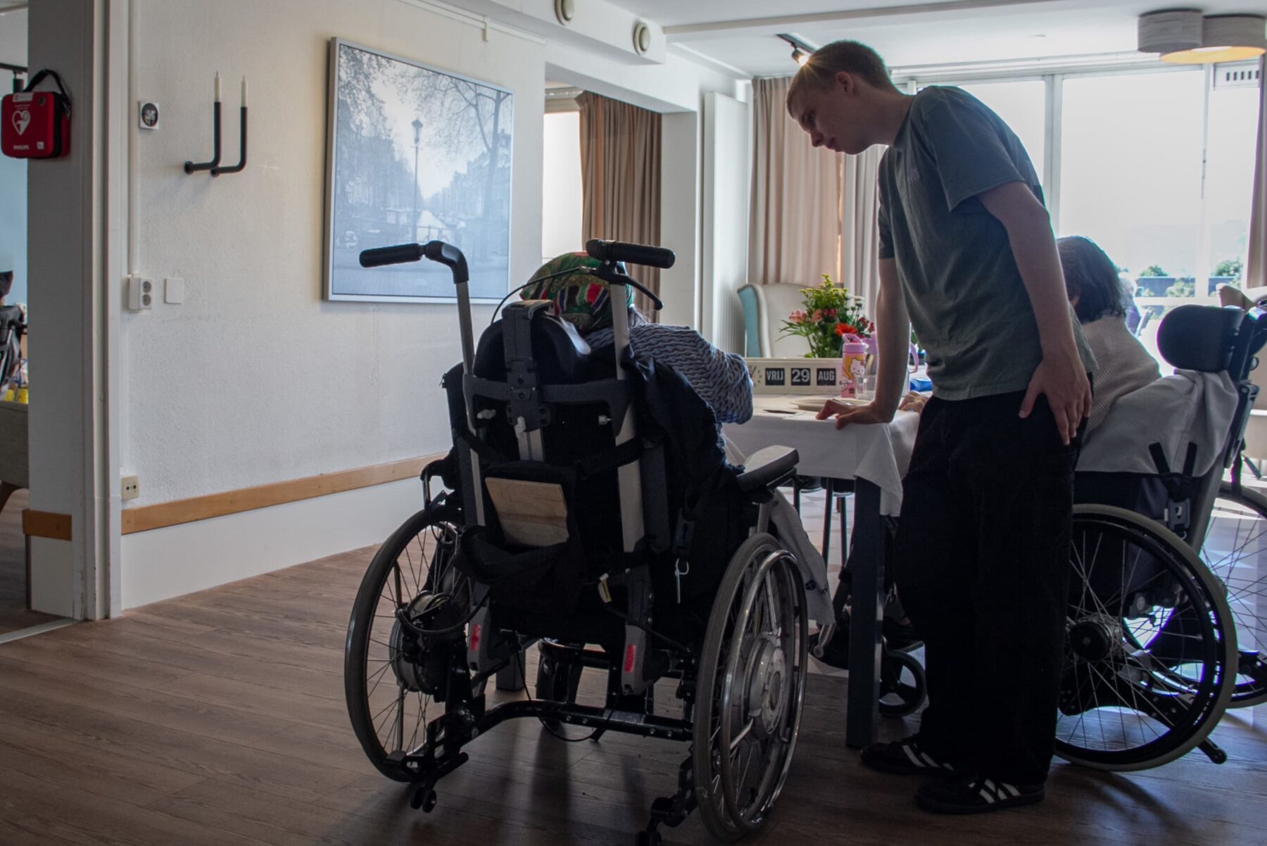 Freiwilliger beugt sich über Tisch, spricht mit Senioren im Rollstuhl im hellen Aufenthaltsraum des jüdischen Altenheims.