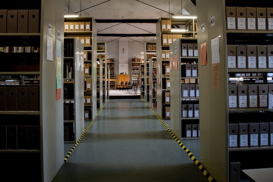 Gang im Staatsarchiv Eupen mit hohen Regalen voller Archivkartons, gelb-schwarze Markierung, Stühle und Kreuz an der Rückwand.