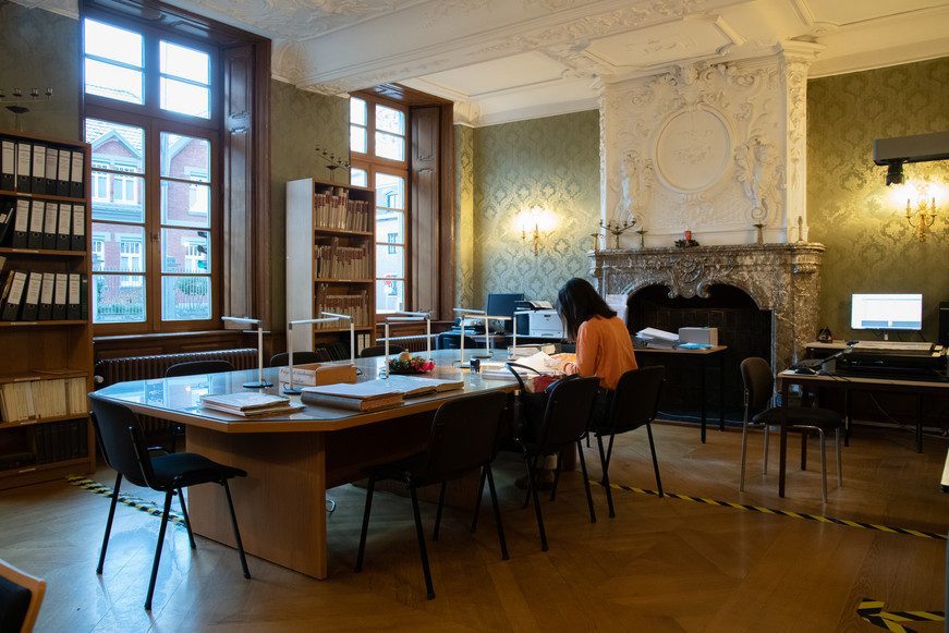 Freiwillige in orange sitzt an Lesetisch im prunkvollen Lesesaal des Staatsarchivs Eupen, große Fenster, Kamin und Regale sichtbar.