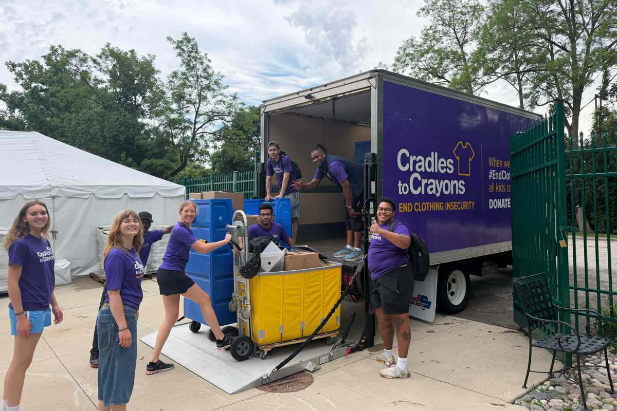 Freiwillige in lila Shirts entladen Spenden aus einem Cradles-to-Crayons-Lkw mit Hebebühne, Wagen und Zelt im Hintergrund.