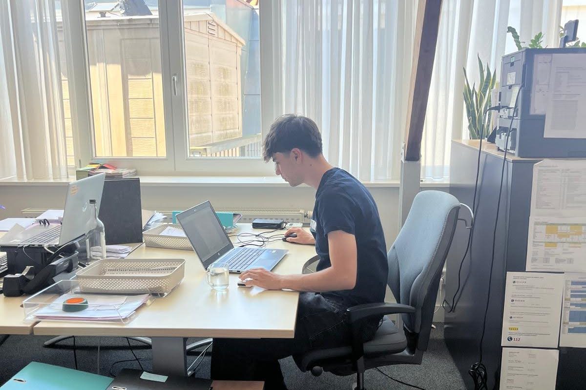 Junger Mann am Schreibtisch in Amsterdamer Büro, arbeitet am Laptop, Papiere und Glas Wasser neben ihm.