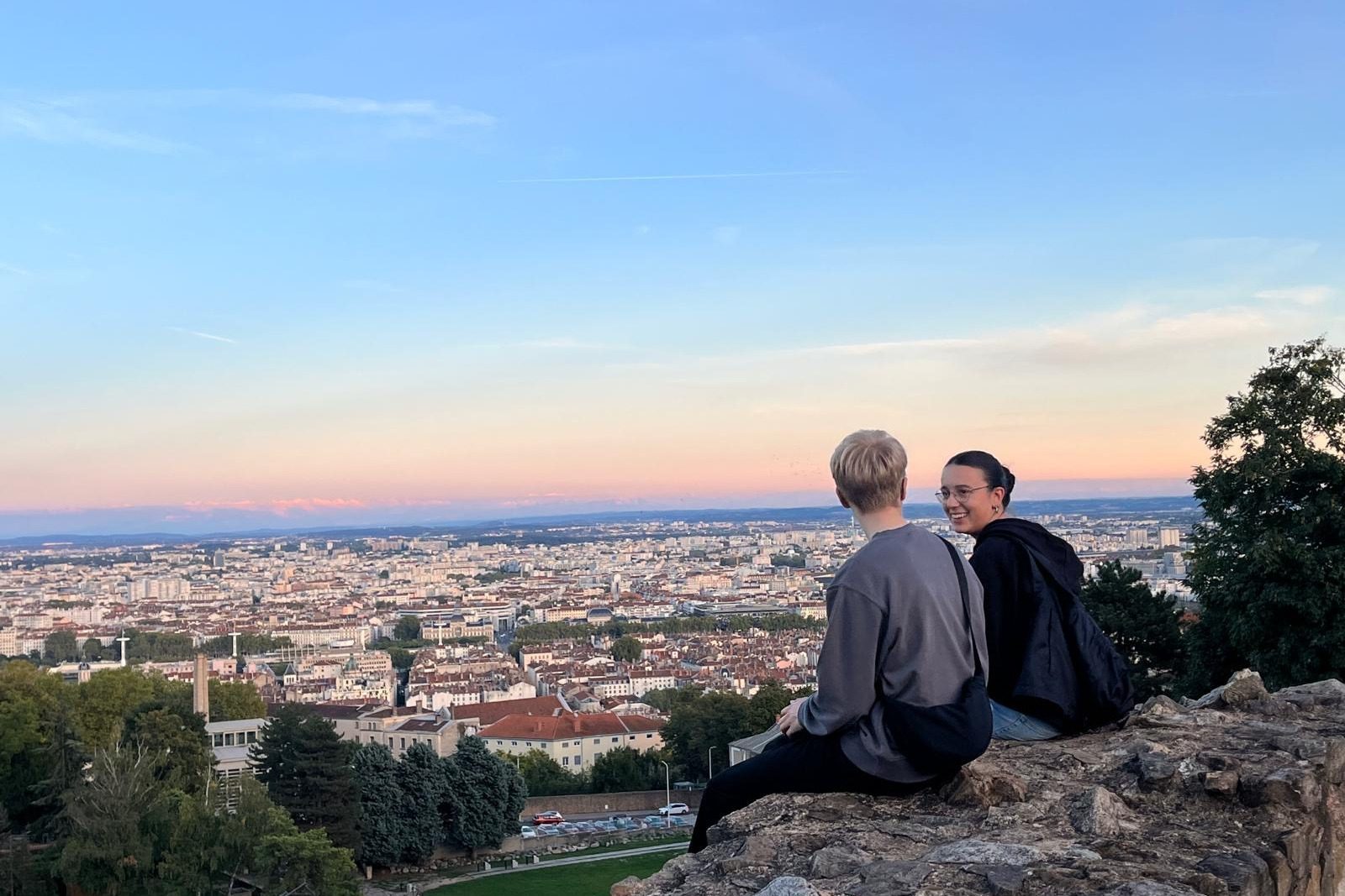 Zwei Freiwillige sitzen auf Ruinen des römischen Theaters und blicken über Lyon im Abendlicht.