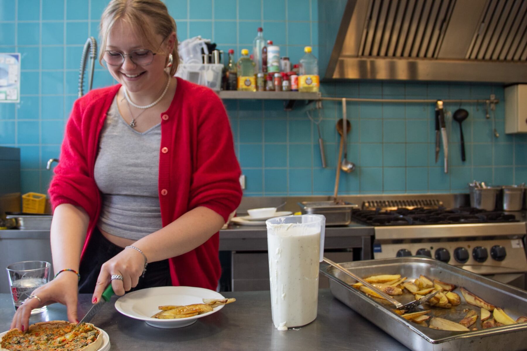 Freiwillige im roten Cardigan schneidet Quiche in Gemeinschaftsküche, daneben Blech mit Kartoffelspalten und Soßkanne.