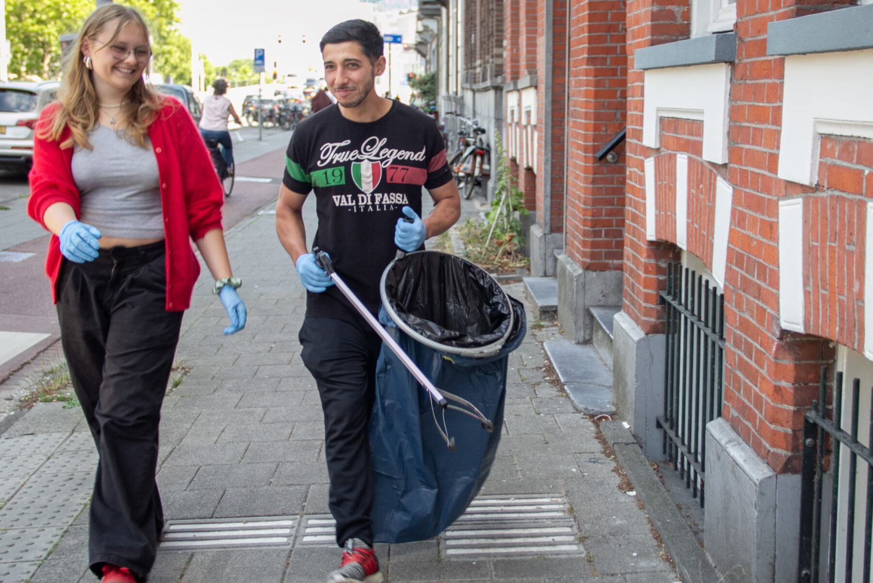 Zwei Freiwillige auf Gehweg in Amsterdam, blaue Handschuhe; Mann trägt großen Müllsack und Greifzange, Frau mit roter Jacke.