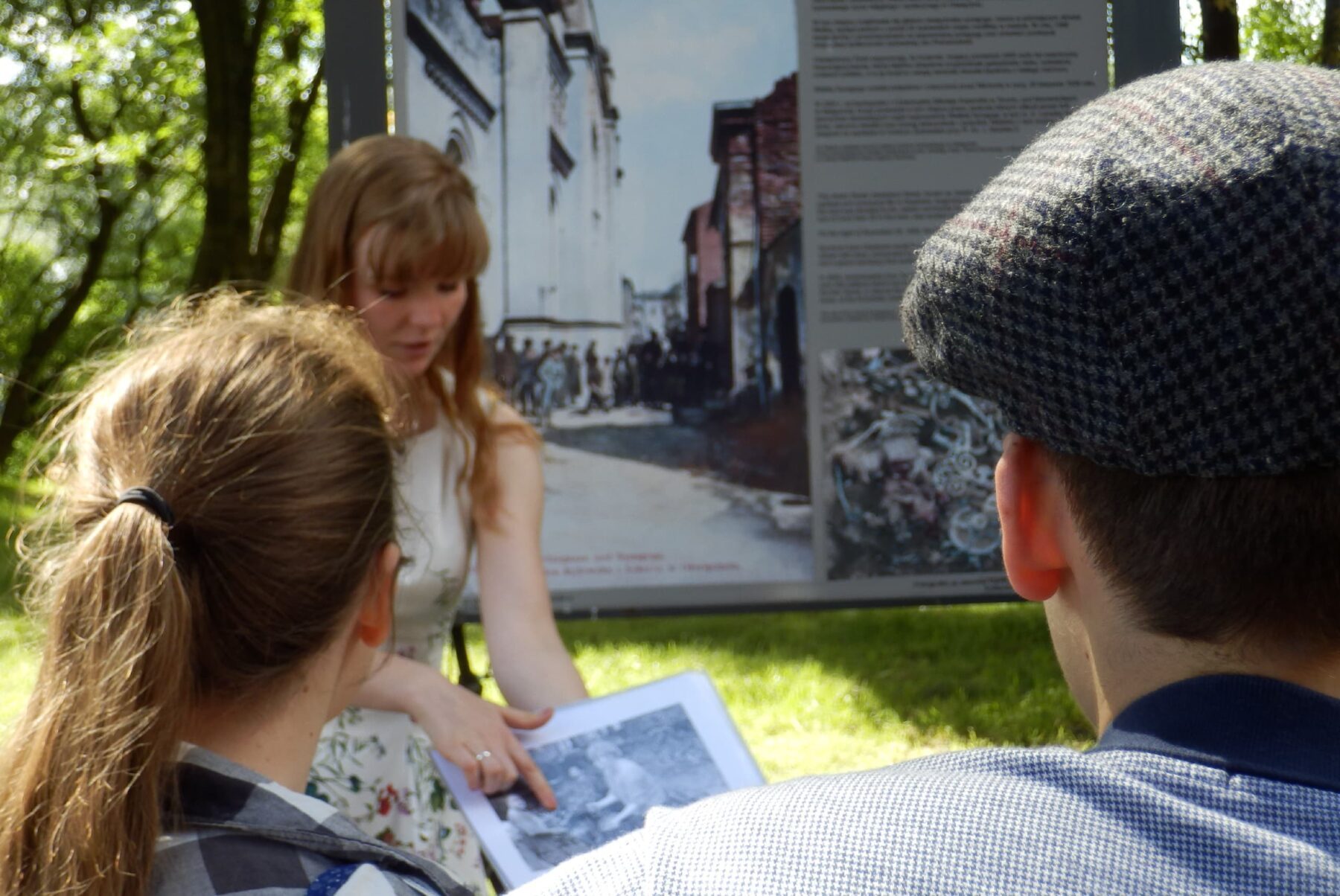 Freiwillige zeigt zwei Zuhörern historische Fotos, deutet auf ausgedrucktes Bild vor großer Infotafel im Park.