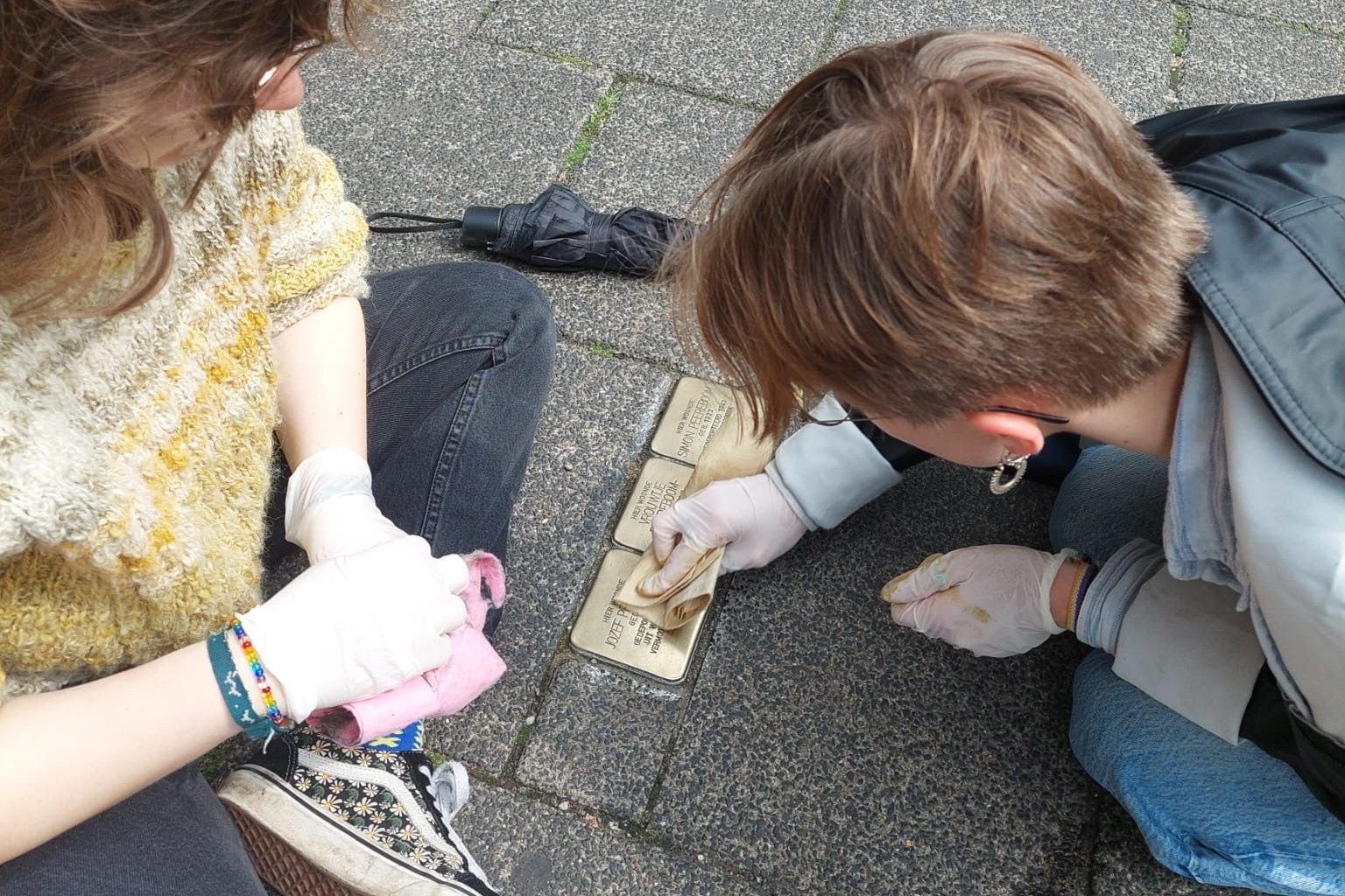 ASF-Freiwillige knien auf Gehweg und polieren mit Handschuhen die Messingtafeln von Stolpersteinen.
