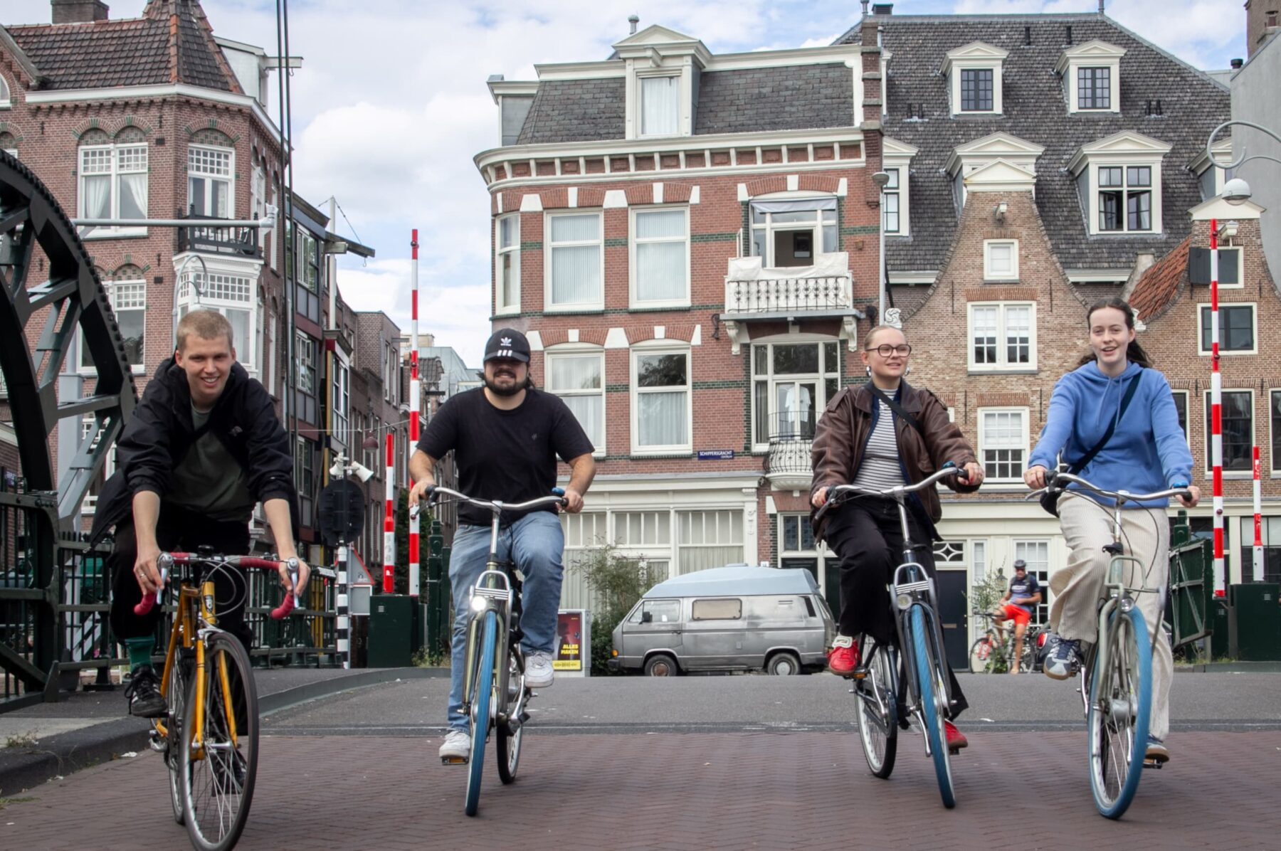 Vier ASF-Freiwillige fahren fröhlich auf Fahrrädern über eine Amsterdamer Brücke vor historischen Backsteinhäusern.