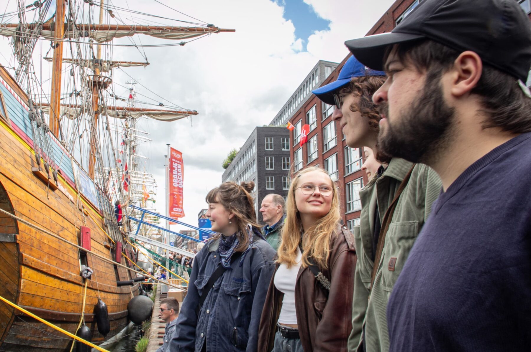 ASF‑Freiwillige am Amsterdamer Kai blicken auf ein großes hölzernes Segelschiff, Gebäude im Hintergrund.
