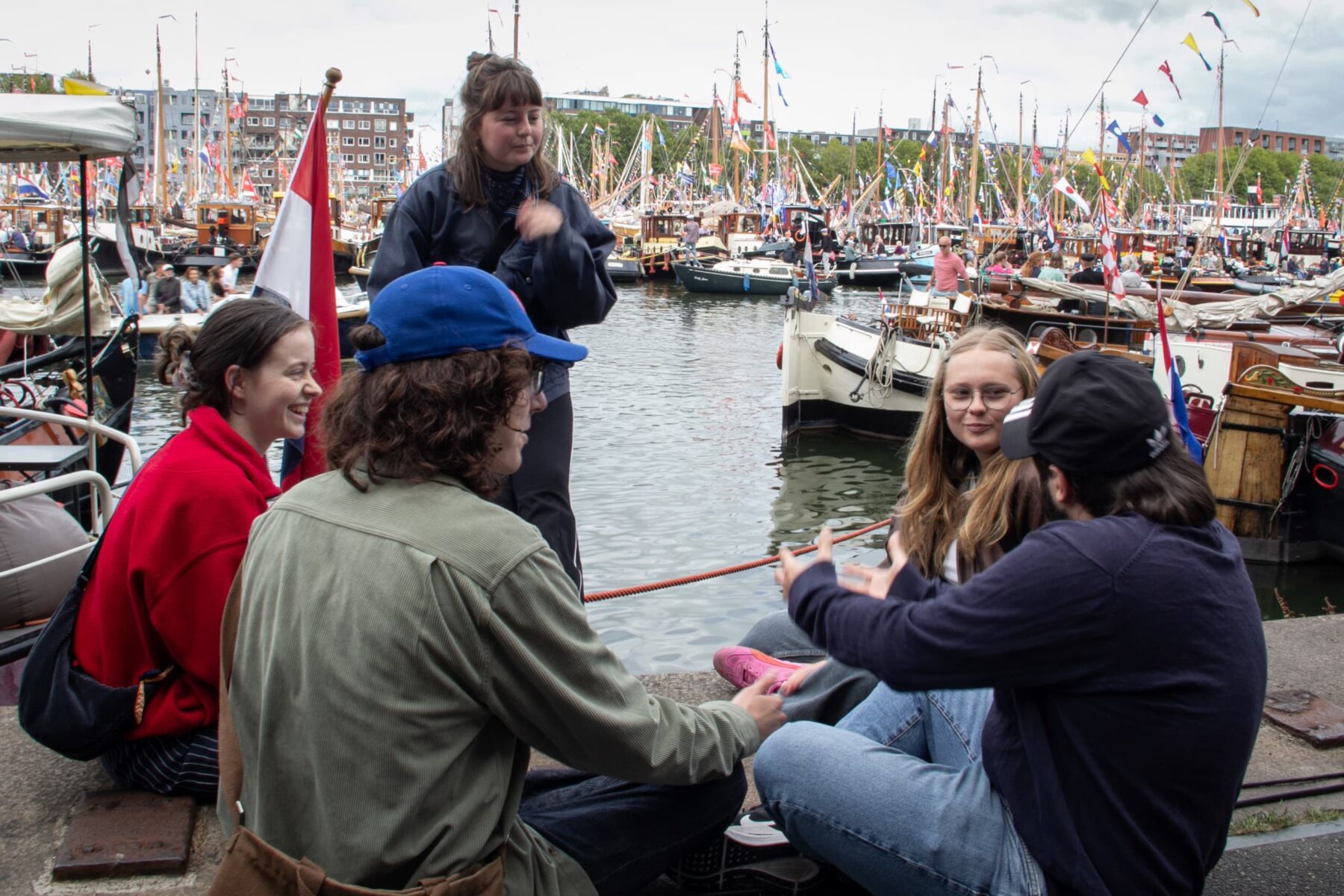 ASF-Ffreiwillige in lockerer Runde am Amsterdamer Hafen, sitzen am Kai vor dicht geflaggten Traditionsseglern