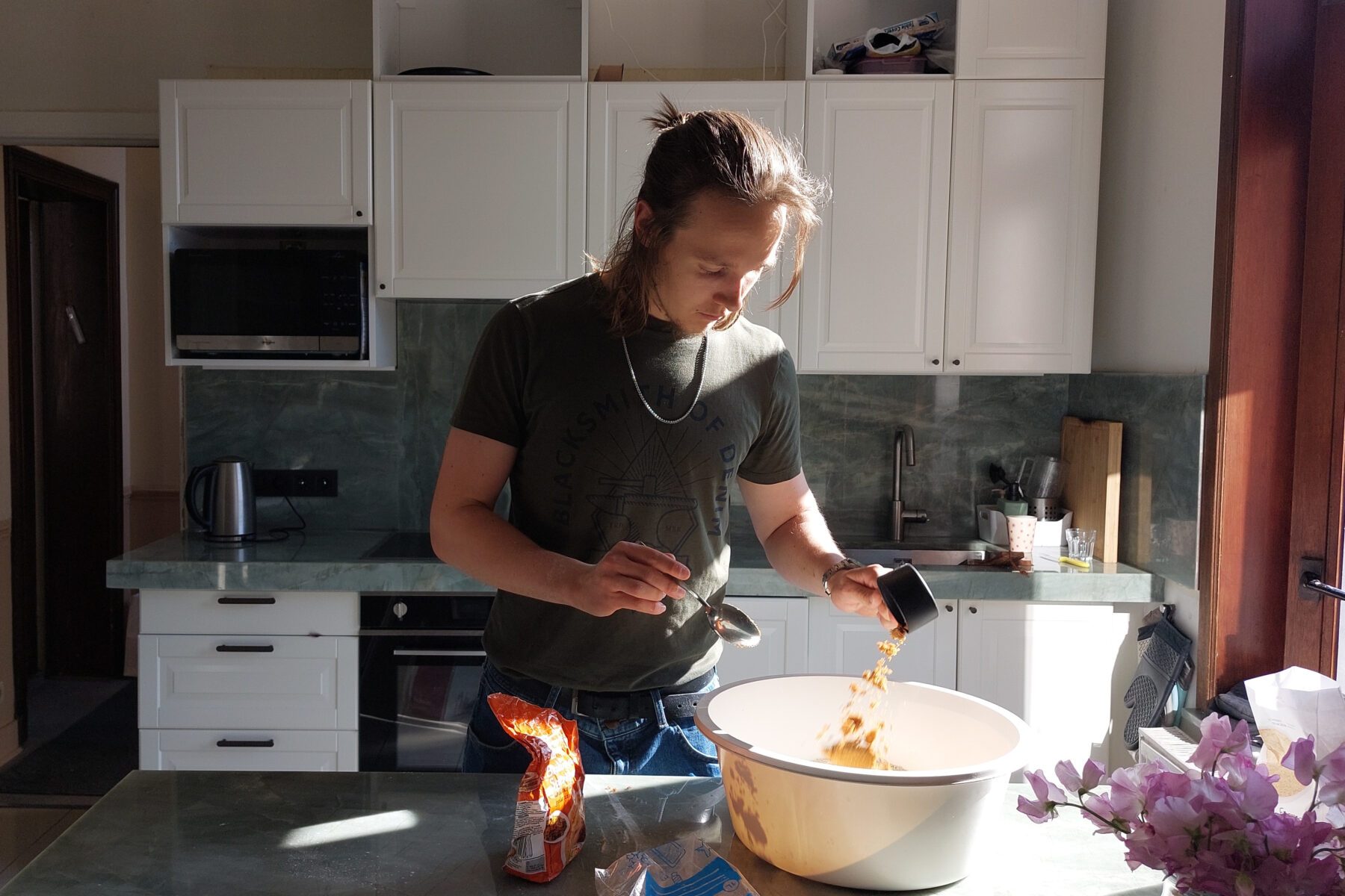 Freiwilliger rührt Zutaten in große Schüssel beim Challabacken in sonniger Küche; lila Blumen am Rand.
