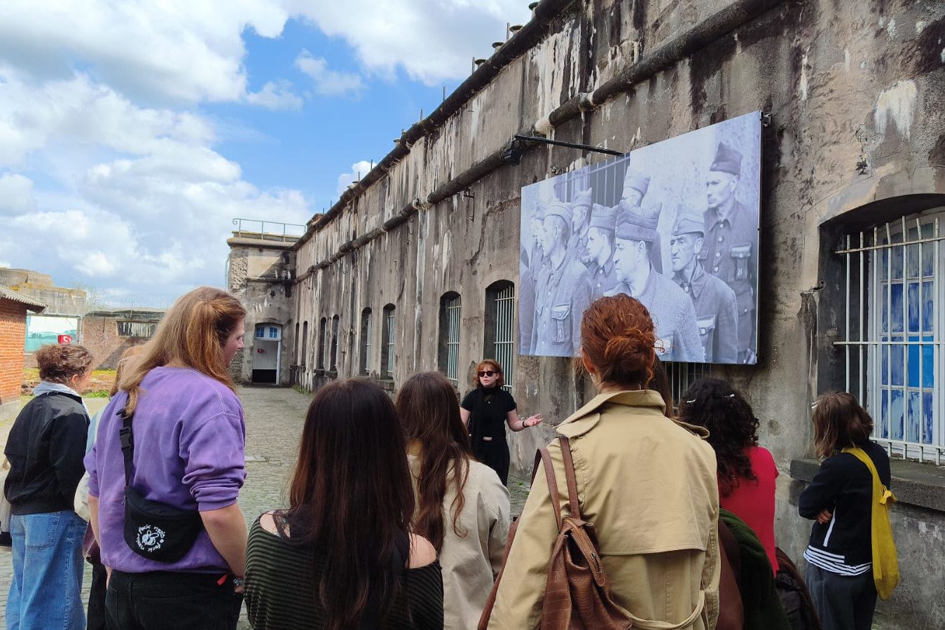 Besuchergruppe bei Führung im Fort Breendonk, Guide gestikuliert vor großer historischer Fototafel.