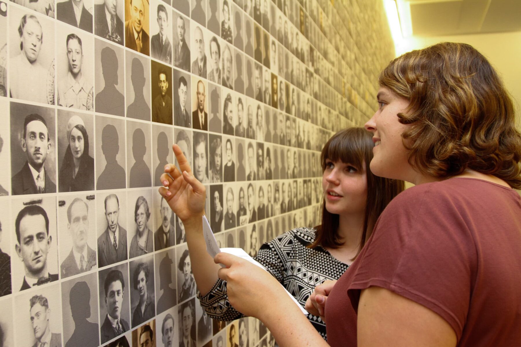 Zwei Personen in der Gedenkstätte Dossin betrachten eine Wand mit Reihen von Porträtfotos; eine zeigt auf ein Bild.