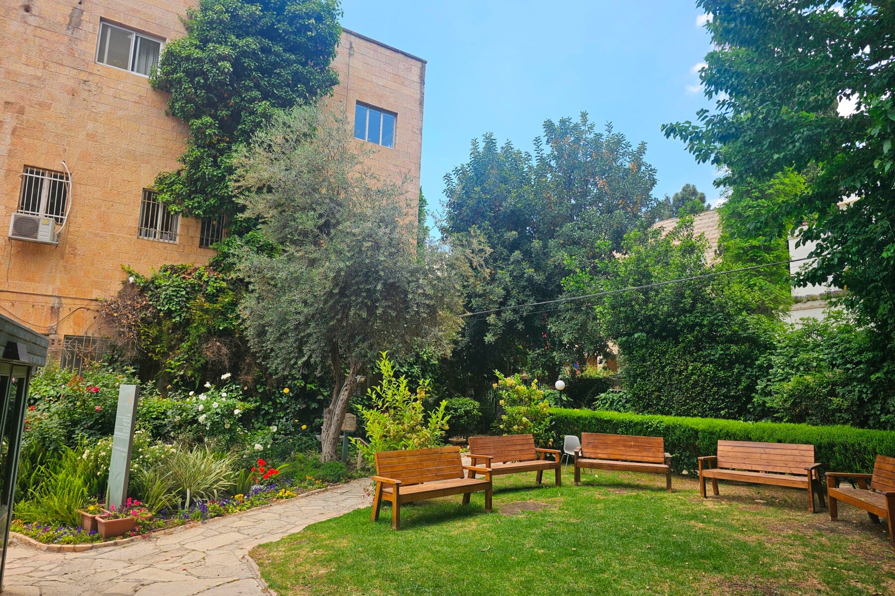 Garten von Yad Ben Zvi in Jerusalem: Rasen mit halbkreisförmigen Holzbänken, Blumenbeeten und Gebäude aus Jerusalemer Stein.