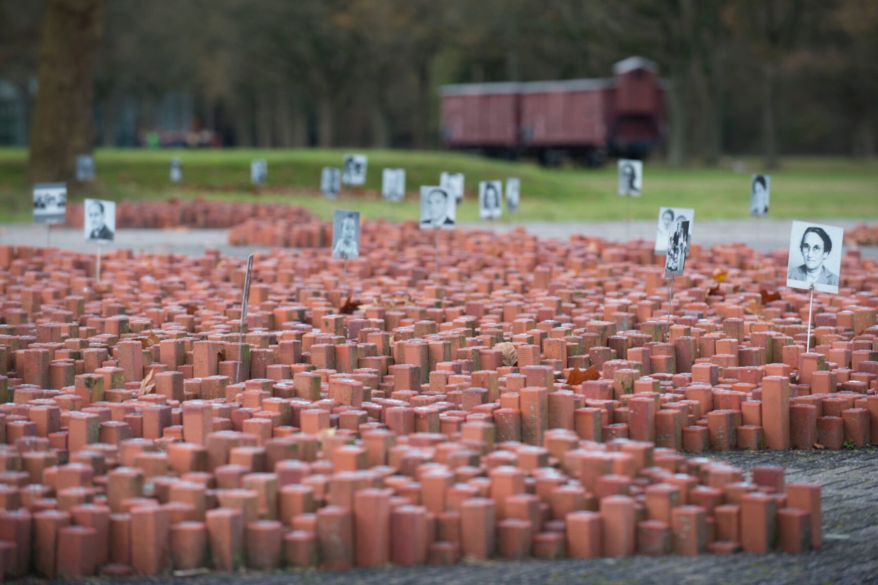 Rote Ziegelstummel und aufgestellte Porträtfotos, im Hintergrund ein Güterwagen auf dem Gedenkort Kamp Westerbork