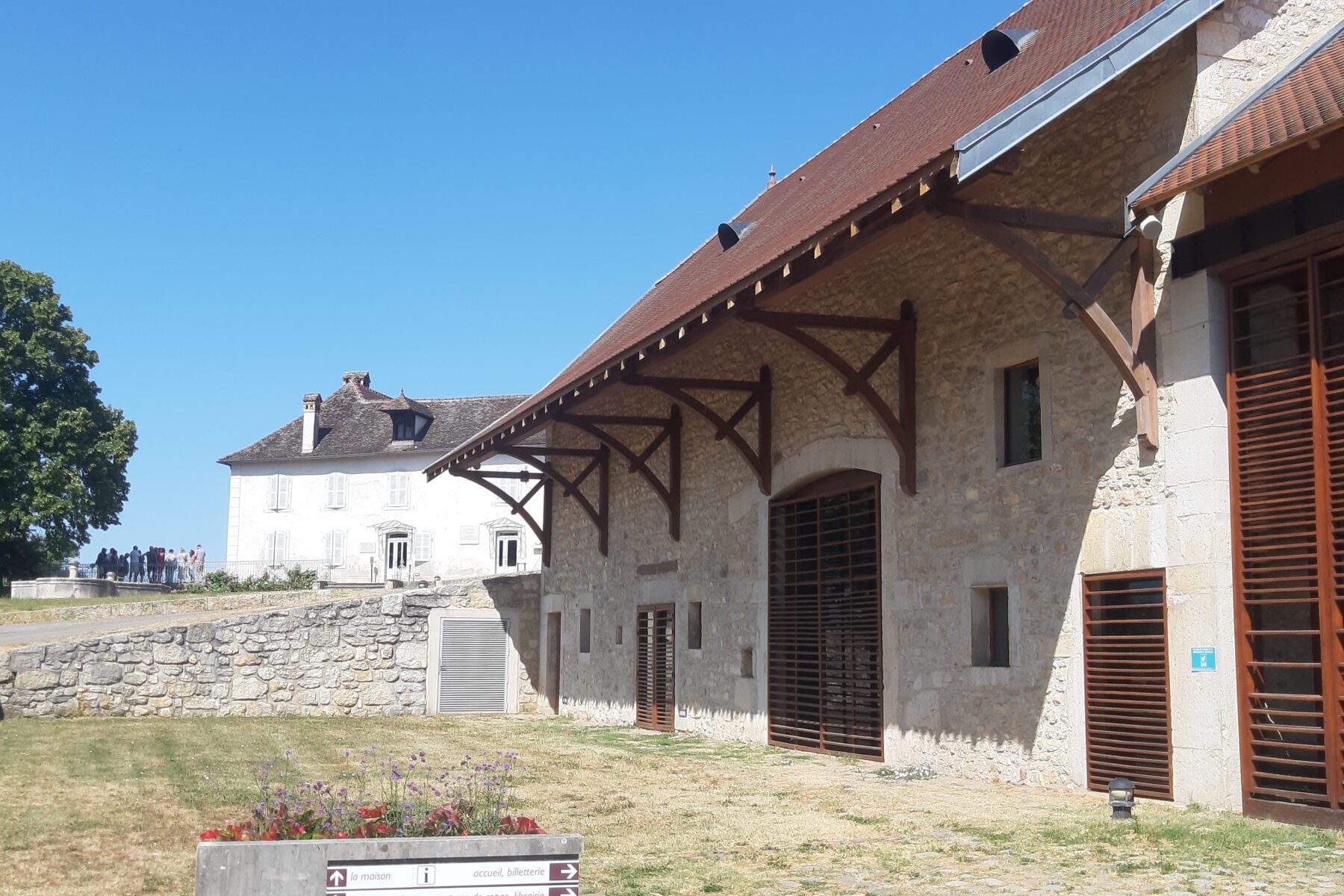 Gedenkstätte Izieu: steinerne Halle mit Holzträgern, Rasen und Hinweisschild vorn; weißes Haus und Besucher hinten.