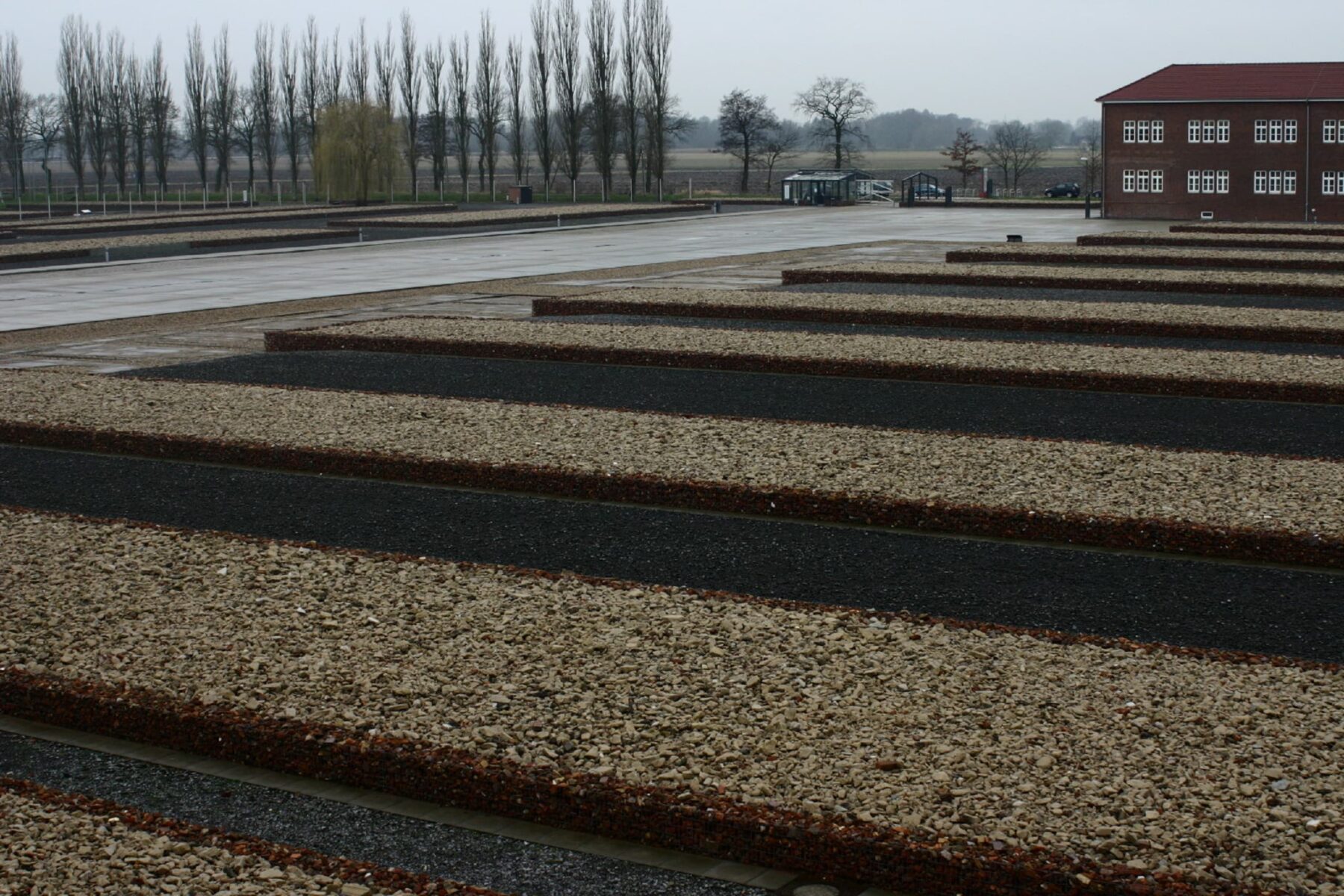 Gedenkstätte Neuengamme: breite, parallele Kies- und dunkle Schotterstreifen vor einem Backsteingebäude bei trübem Himmel