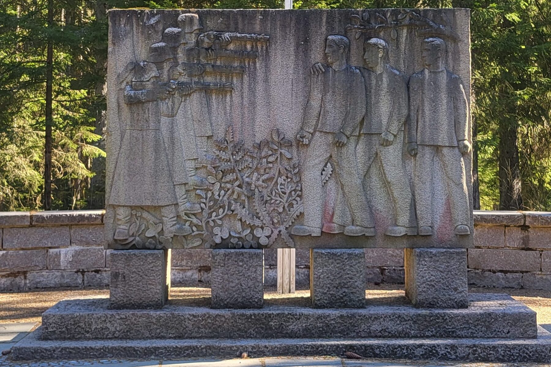 Steinrelief der Falstad-Gedenktafel: Soldaten richten Gewehre auf drei gefesselte Männer, Pflanzenmotiv, Wald im Hintergrund.