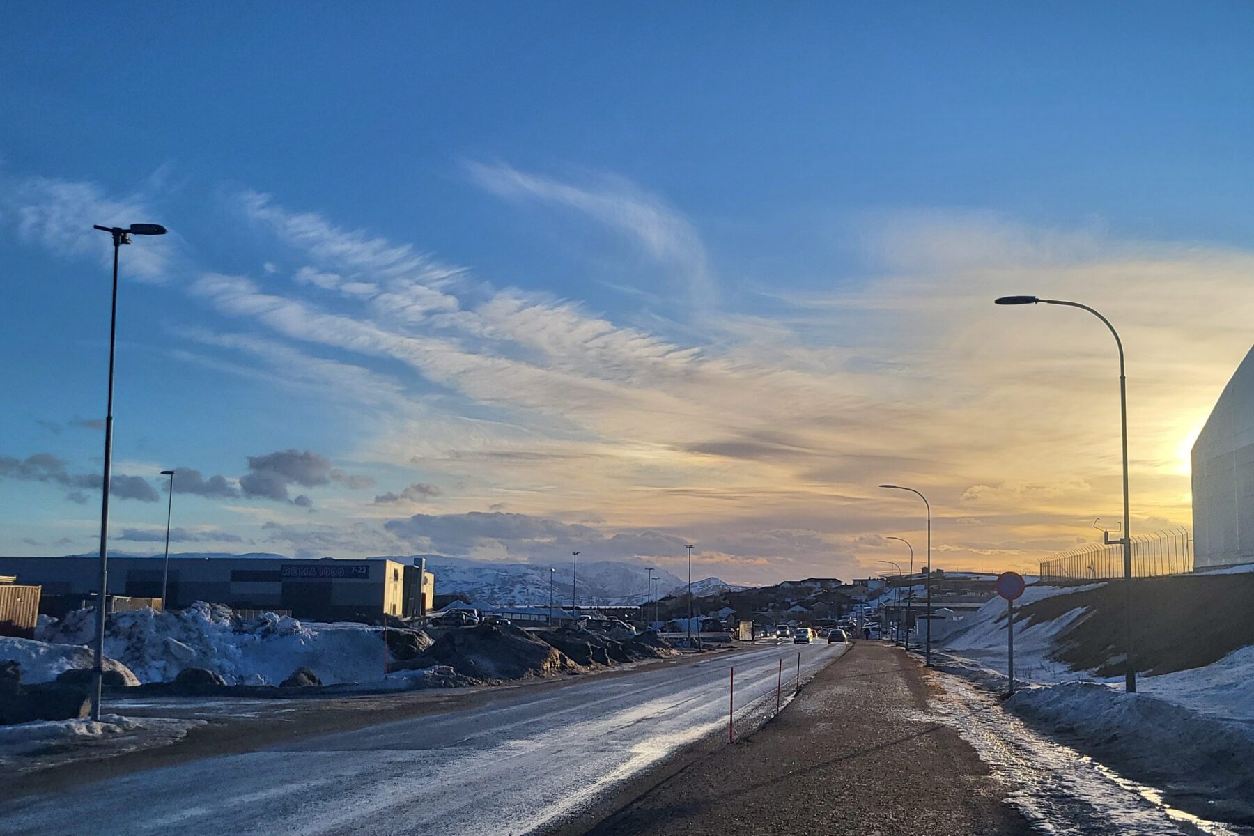 Straße in Hammerfest, Norwegen bei Sonnenuntergang; nasse Fahrbahn, Schneeberge, Laternen und entfernte Berge.