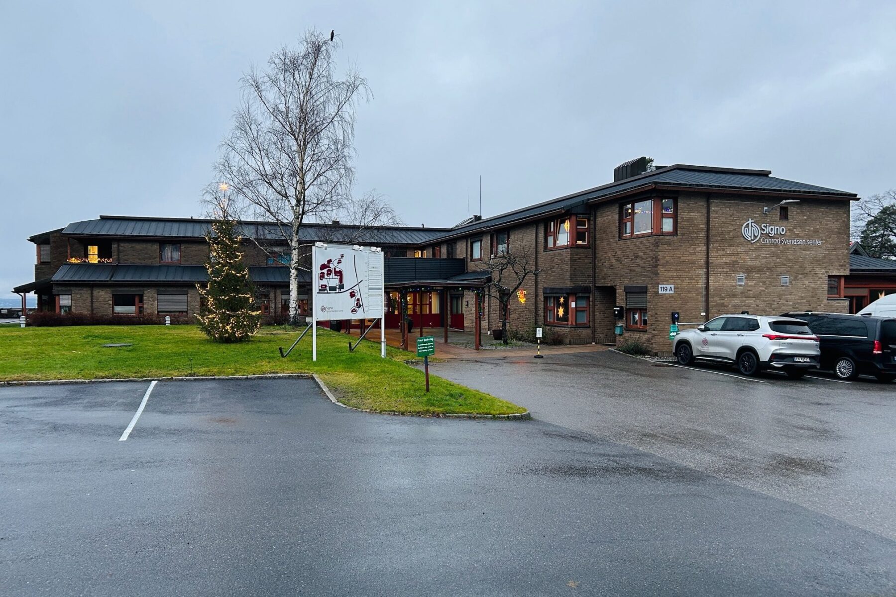 Signo-Hauptgebäude in Oslo: zweistöckiges Backsteingebäude, beleuchteter Weihnachtsbaum, nasser Parkplatz und parkende Autos.