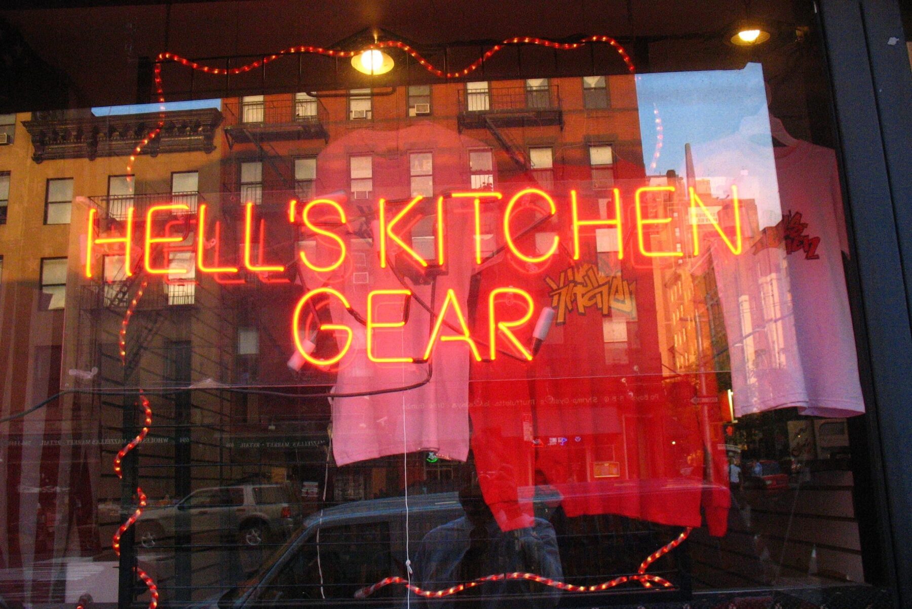Neonschild HELL'S KITCHEN GEAR im Schaufenster, rote/pinke Shirts, Spiegelung von Backsteingebäuden und Autos