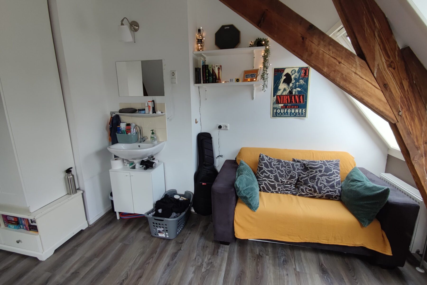 Dachzimmer mit gelbem Sofabezug, gemusterten und grünen Kissen, Waschbecken, Gitarrenkoffer, Nirvana-Poster.