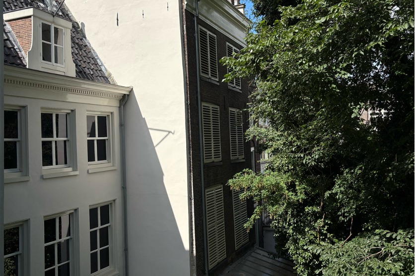 Hinterhaus des Anne-Frank-Hauses in Amsterdam: weiße Wand, dunkles Backsteinhaus mit Fensterläden, Sonnenschatten und Bäume.