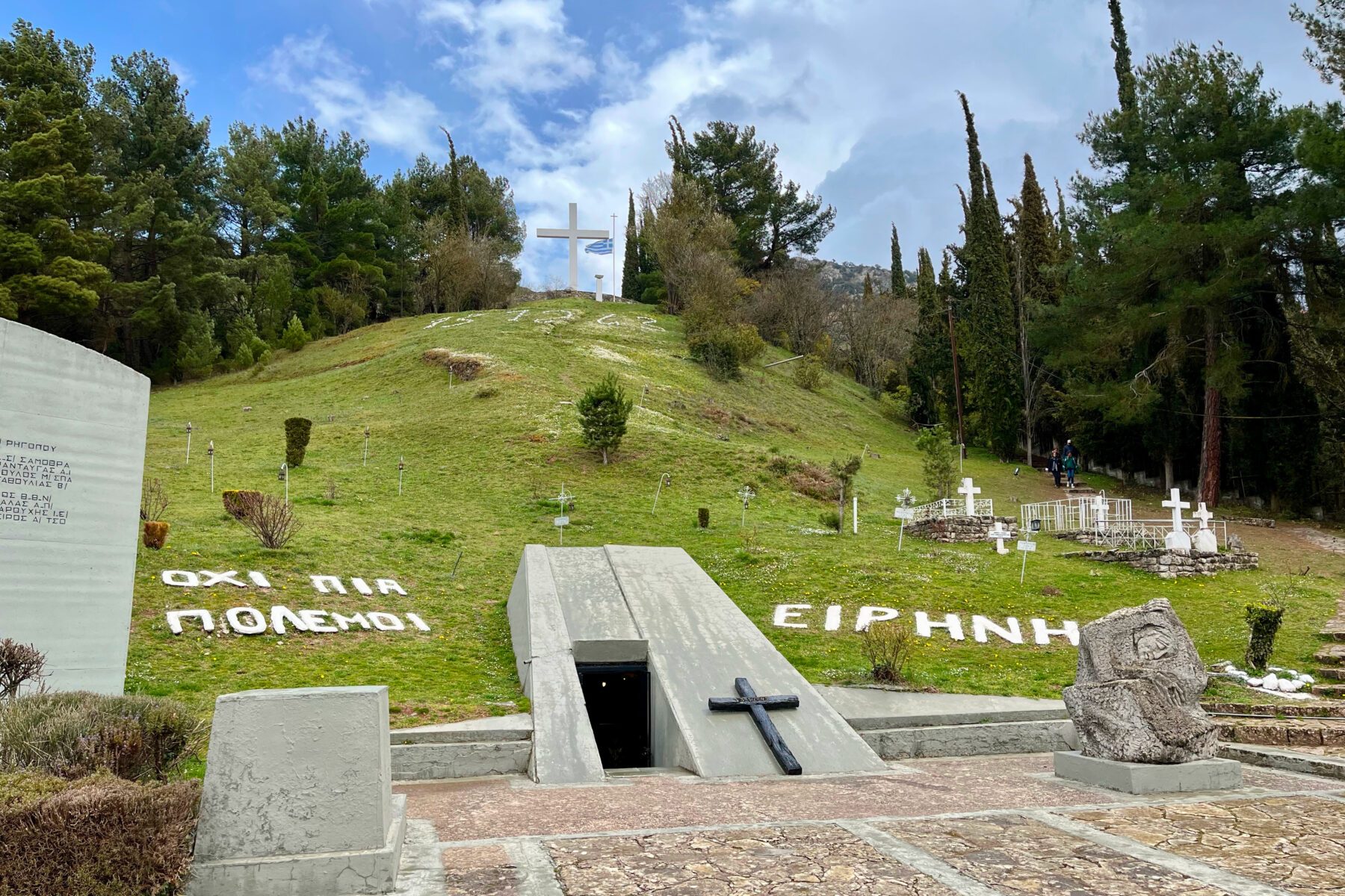Holocaust-Gedenkstätte Kalavryta: Hügel mit großem weißem Kreuz, Betoneingang, viele Kreuze und Schriftzug ‚EIRHNH‘.