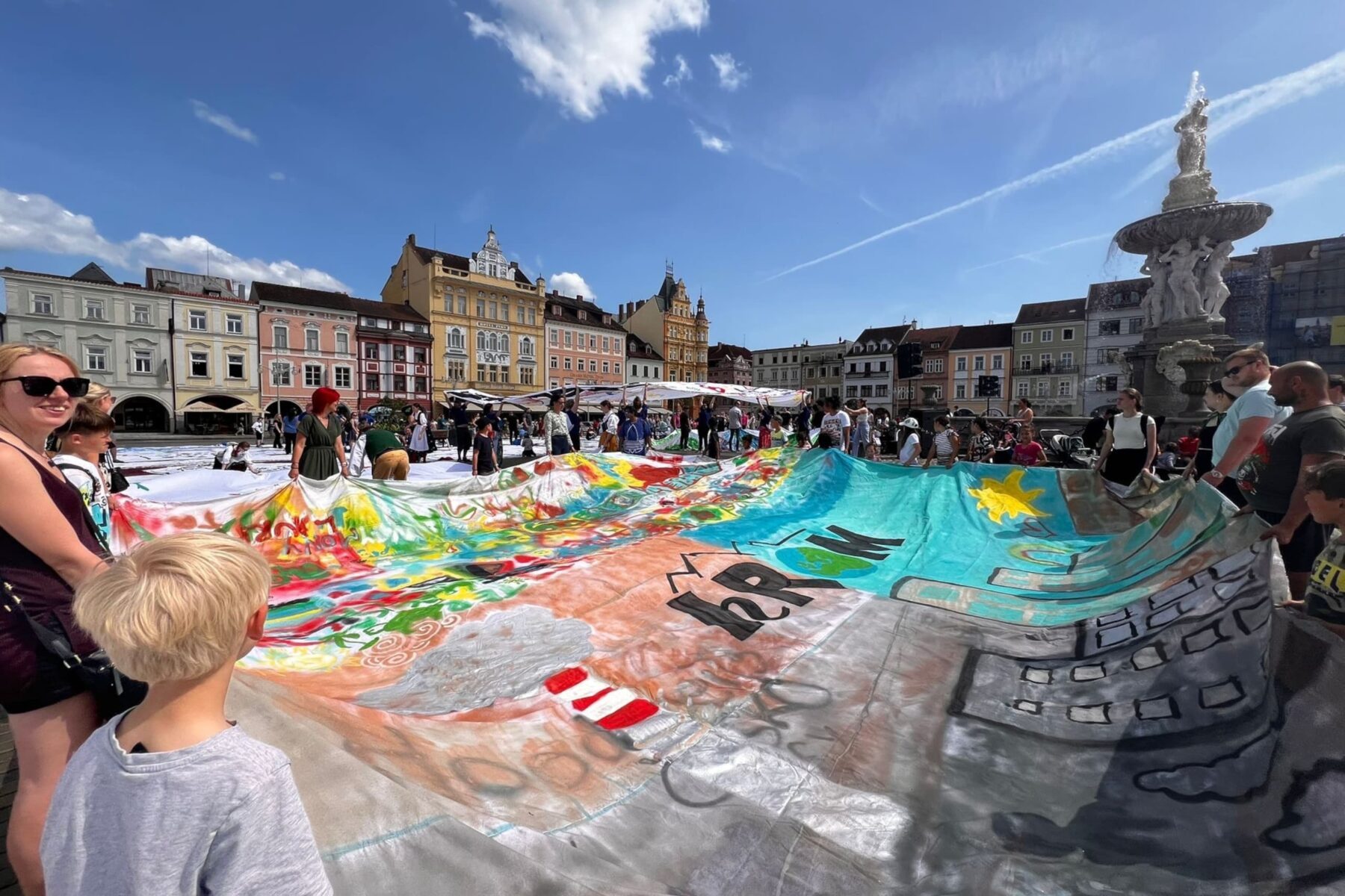 Riesiges bunt bemaltes Transparent, von Menschen auf historischem Marktplatz mit pastellfarbenen Häusern und Brunnen gehalten.
