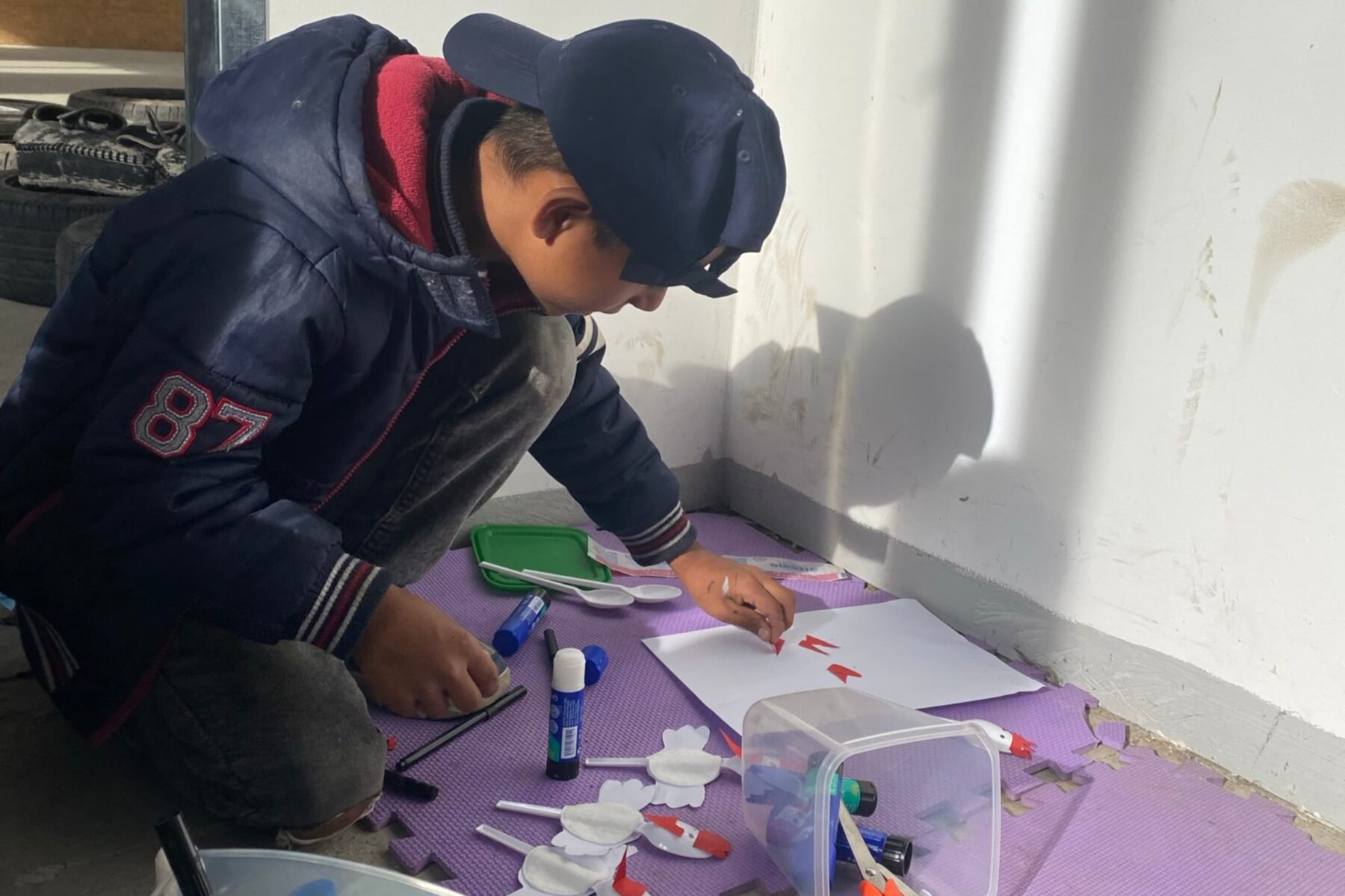 Junge bei Bastelarbeit auf lila Matte, klebt rote Papierstücke, umgeben von Klebestiften, Schere und Plastikbehälter.