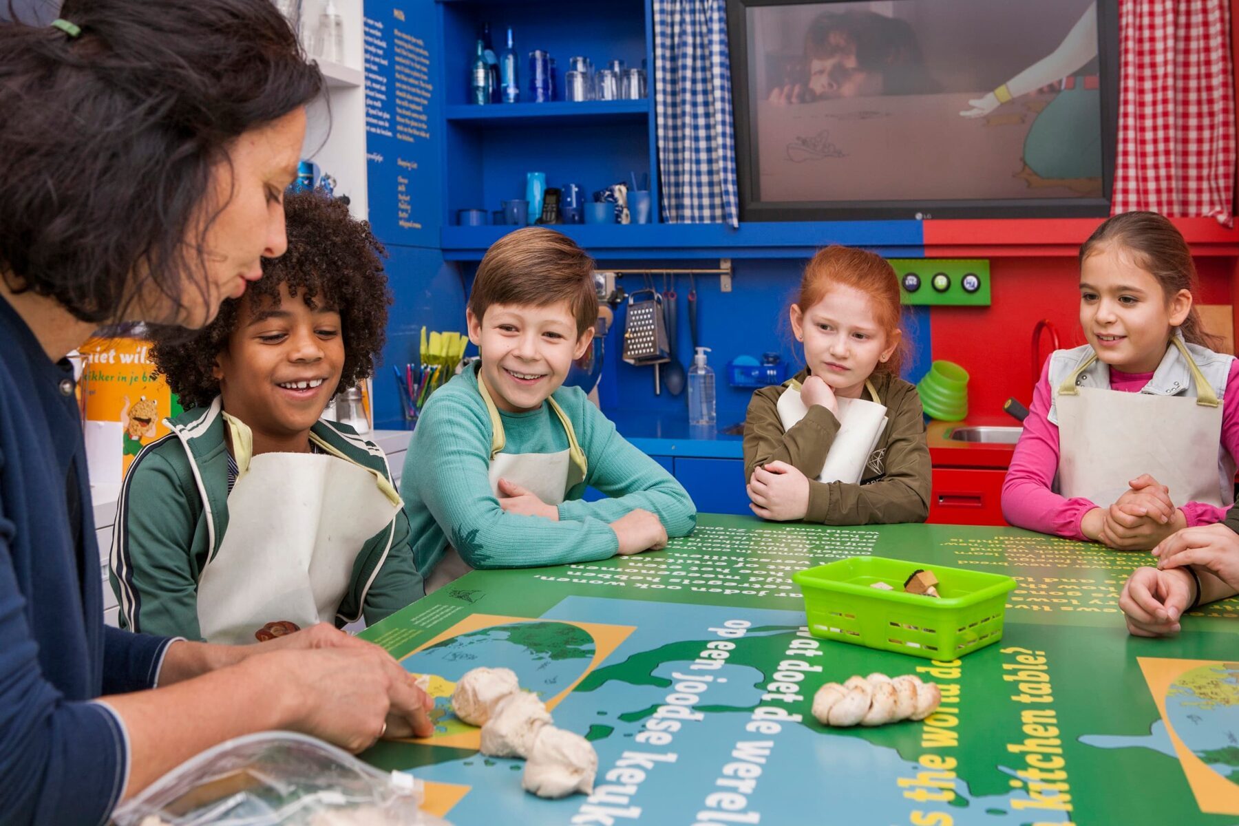 Kochkurs im Kindermuseum des Jüdischen Museums Amsterdam: Leiterin formt Teig, vier Kinder mit Schürzen am bunten Tisch.