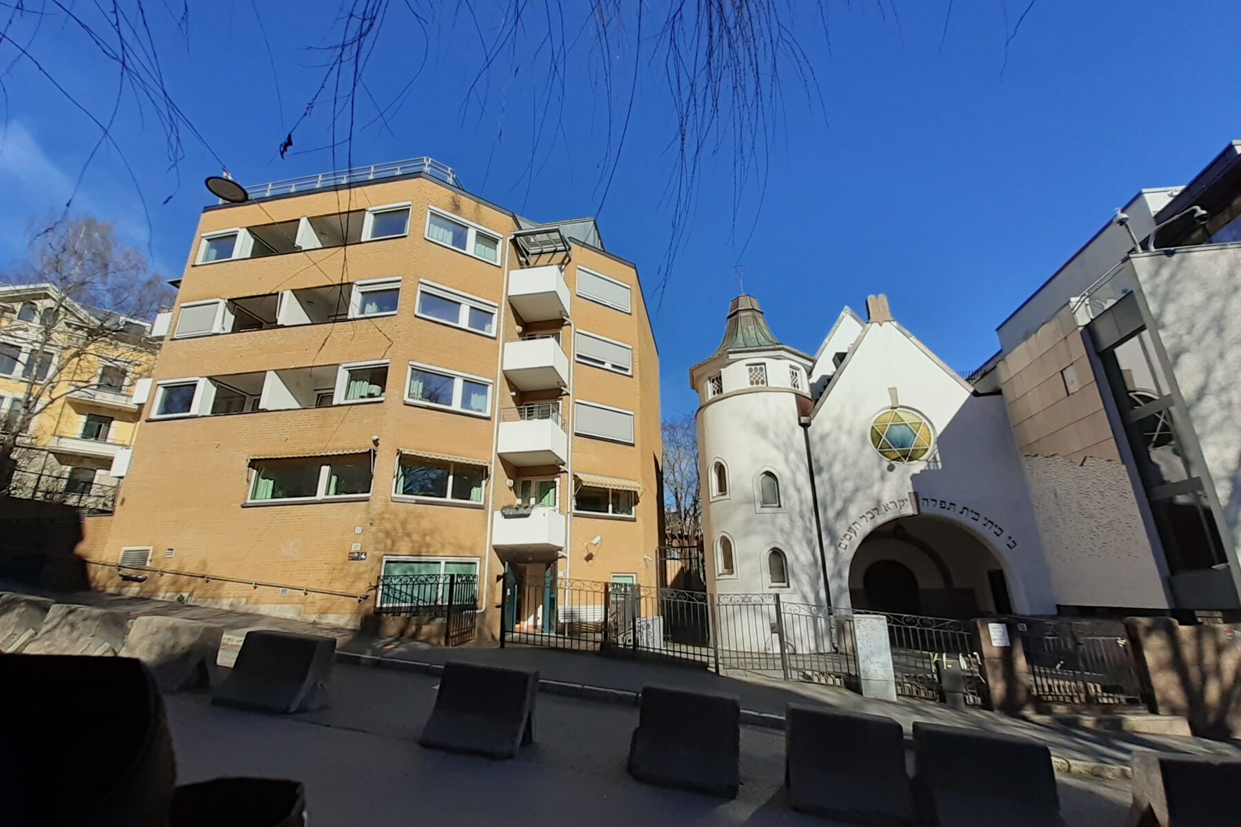 Beige jüdisches Seniorenwohnheim links, weiße Synagoge mit Davidstern und Rundturm rechts, Gittertor, blauer Himmel.