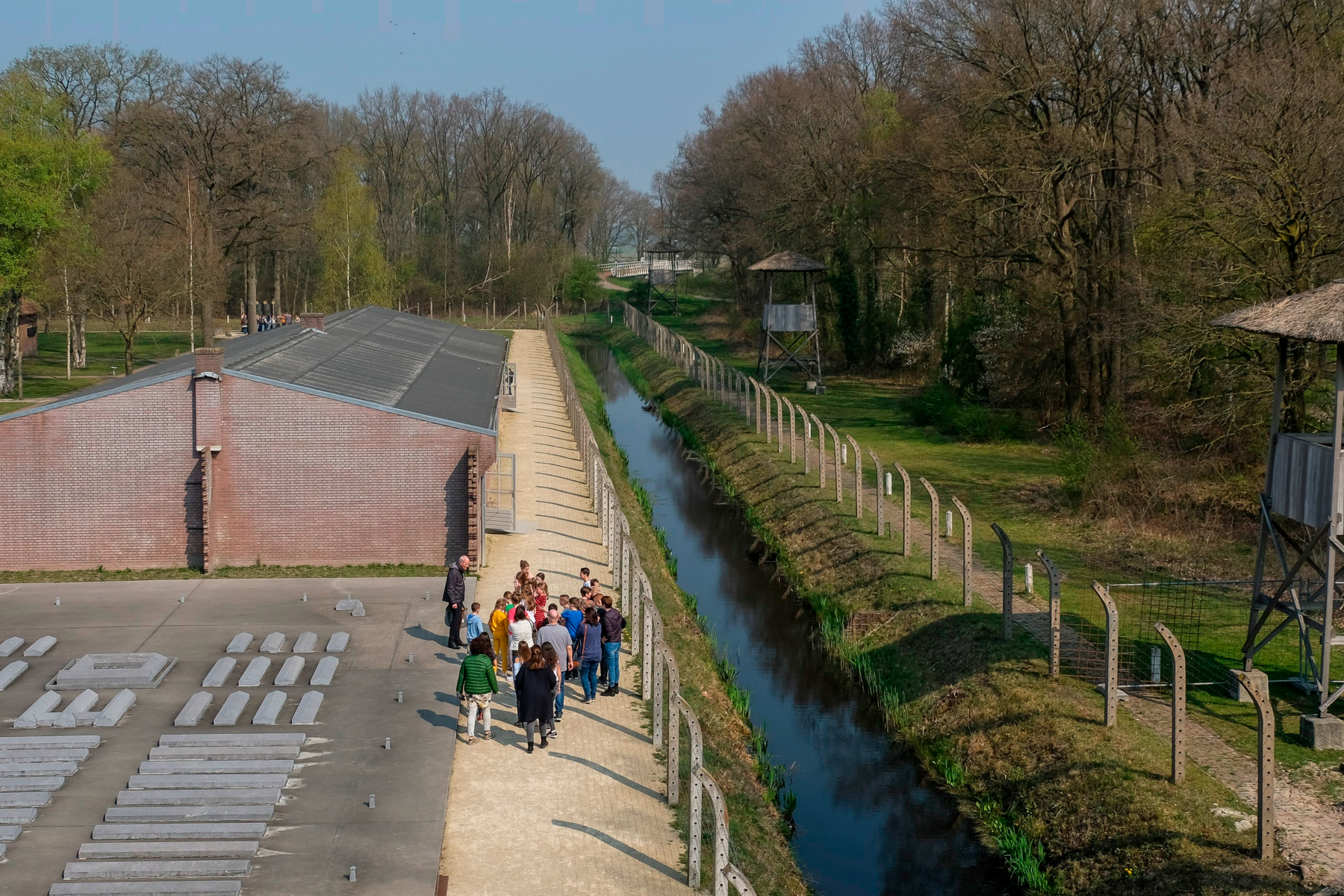 Gruppenführung im Kamp Vught (Niederlande) neben Backsteinbaracke, Wassergraben und Wachtürmen, von oben