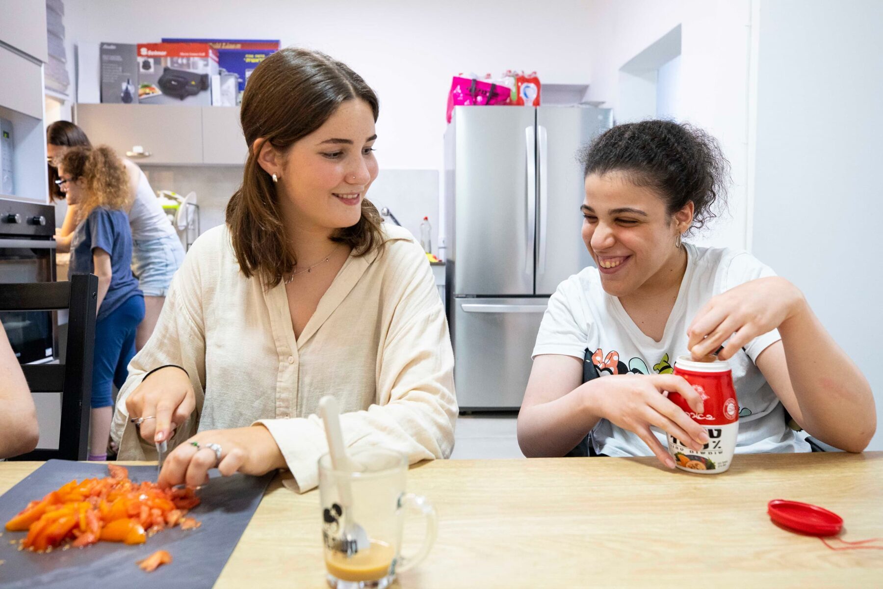 Zwei junge Frauen in Küche: links schneidet Tomaten, rechts lacht und öffnet ein Glas.