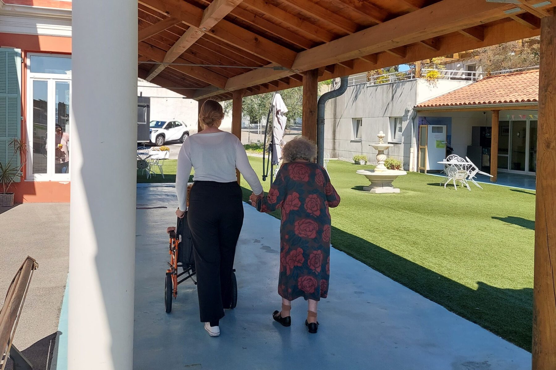 Pflegekraft schiebt Rollstuhl, hält Hand einer älteren Bewohnerin auf überdachtem Weg im Hof von La Colline (Nizza)
